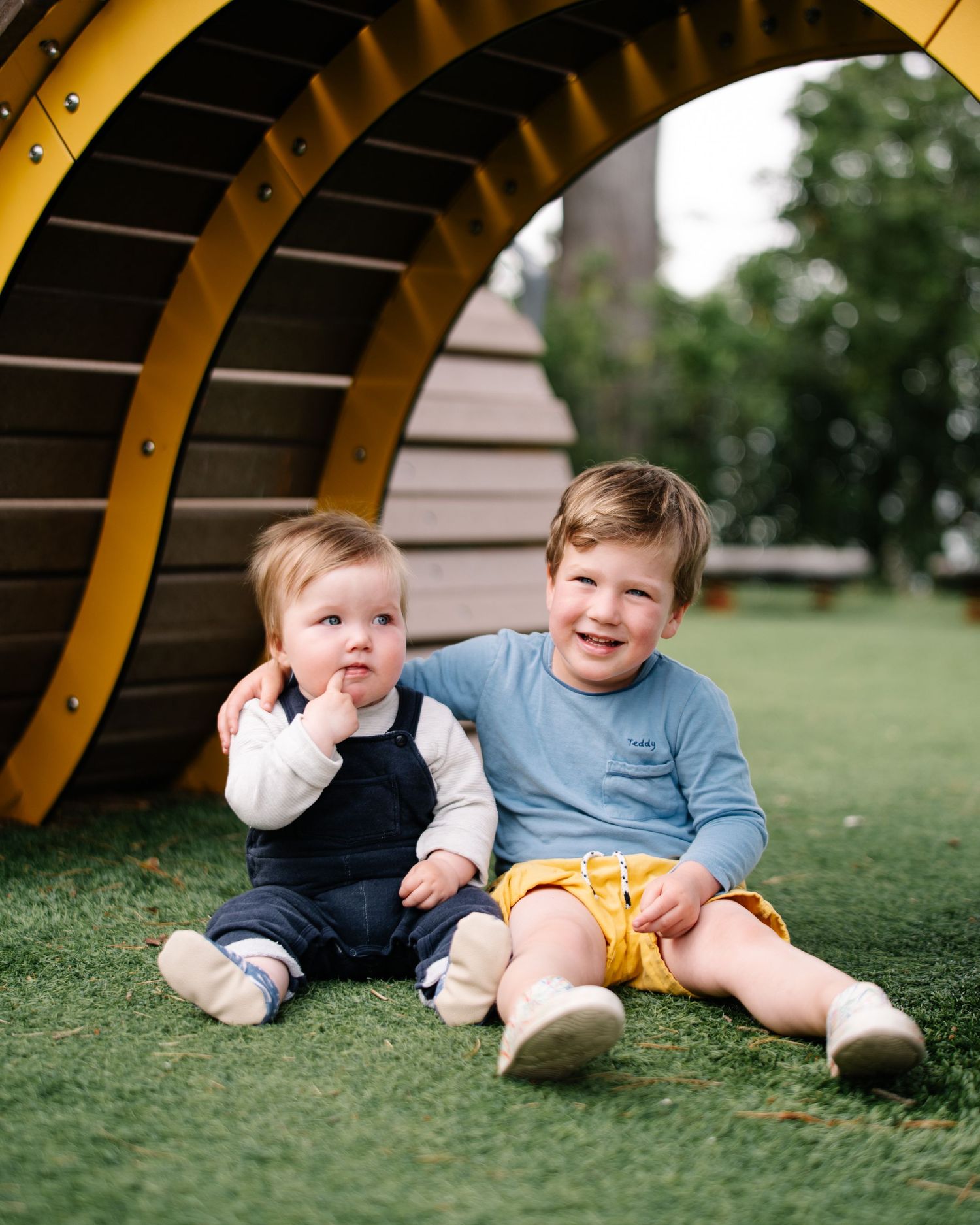 Two young siblings sit smiling on green grass beside a wooden playground structure outdoors.