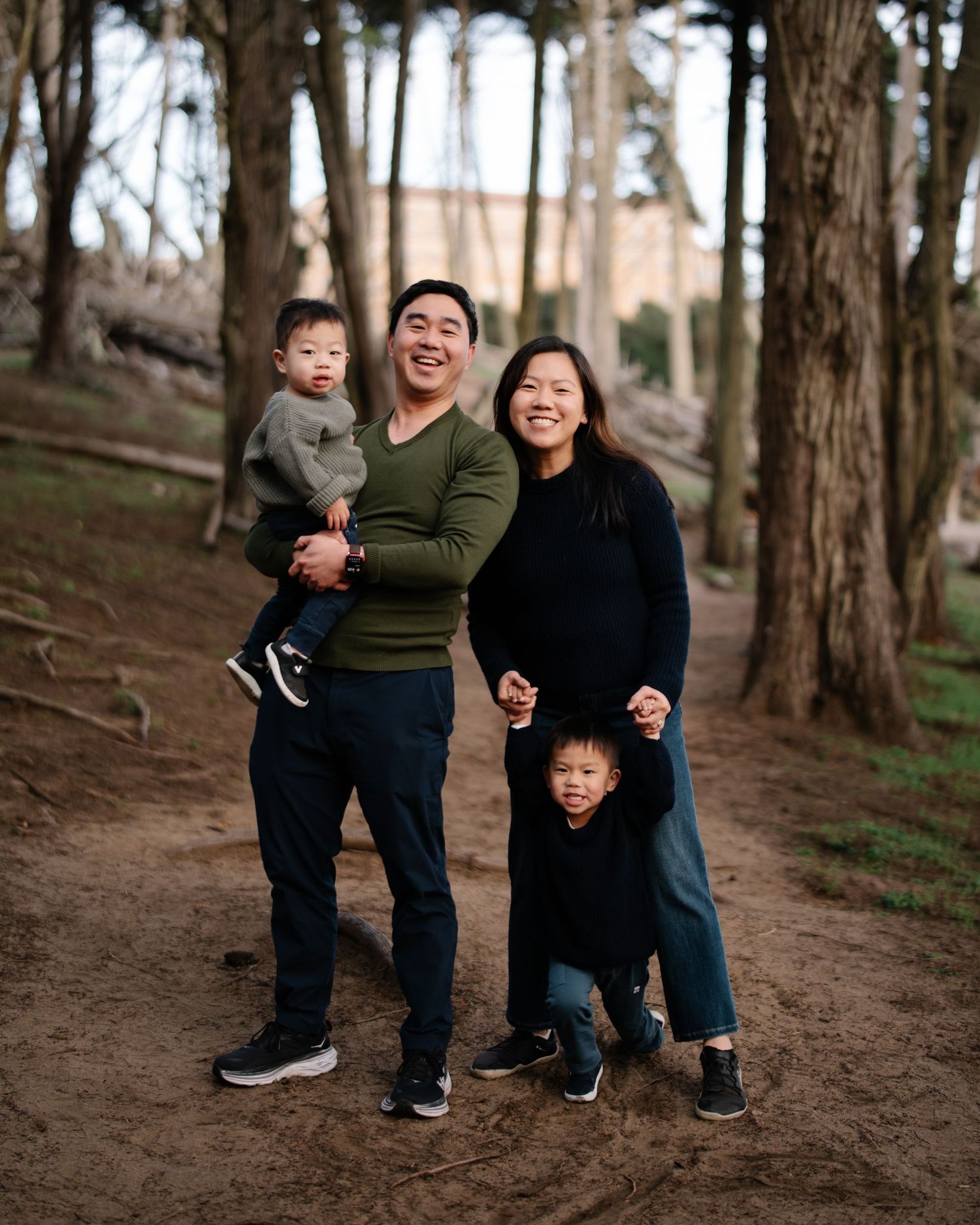 A smiling Asian family of four poses outdoors among trees, with dad holding a toddler and mom beside an older child.