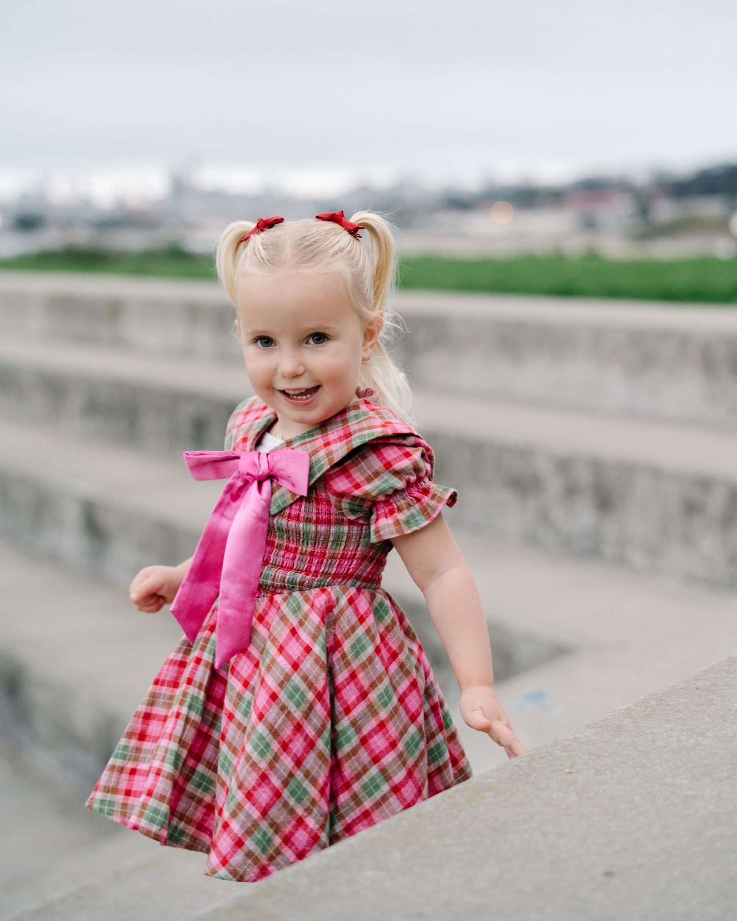 A young child in pigtails wearing a pink and plaid dress poses outdoors on concrete steps.