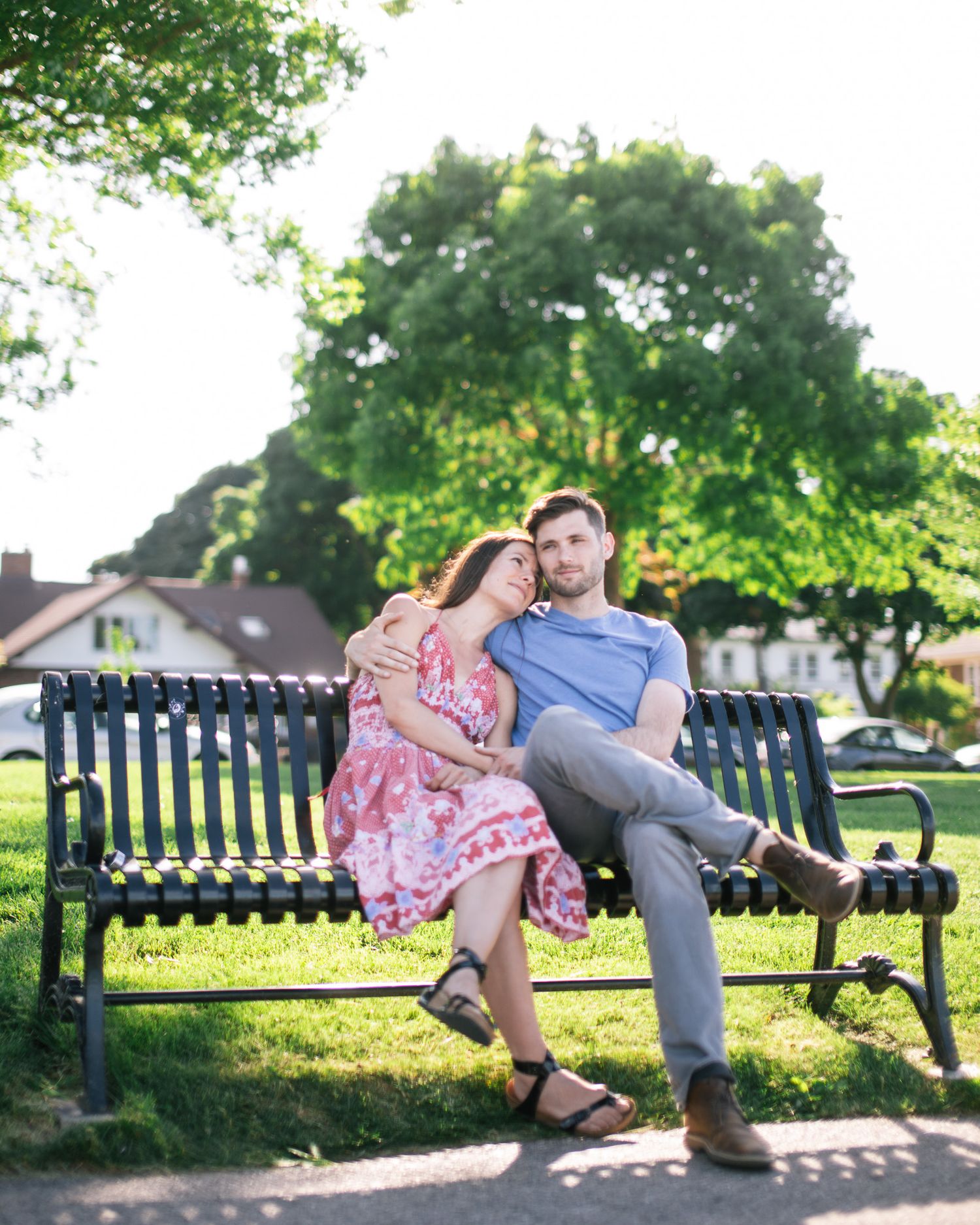 A couple sits together on a park bench on a sunny day with lush green trees in the background.