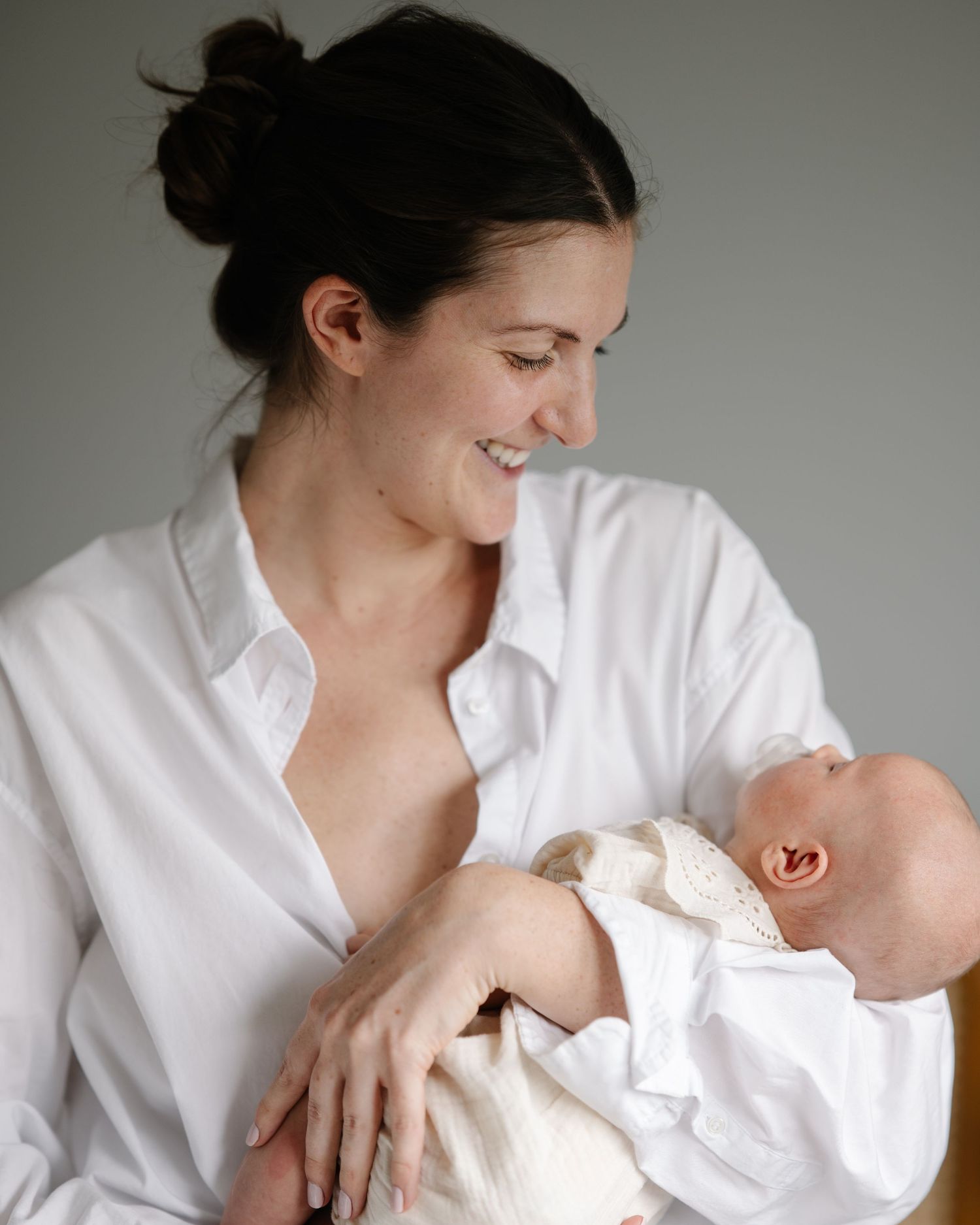 Smiling mother in white shirt breastfeeding her newborn baby, captured in a soft, natural light portrait.