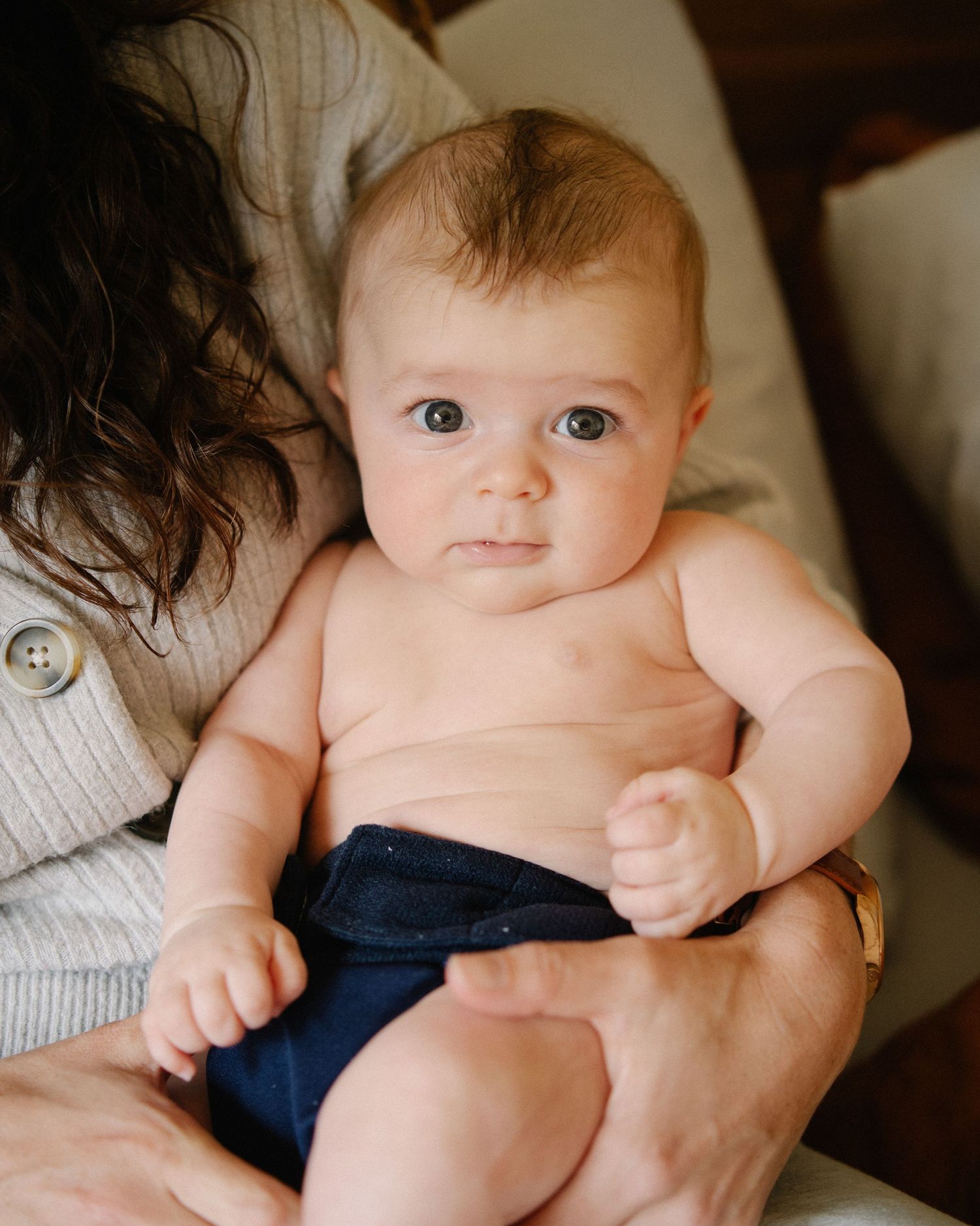 A baby wearing navy blue cloth diaper sits on someone's lap while looking at the camera.