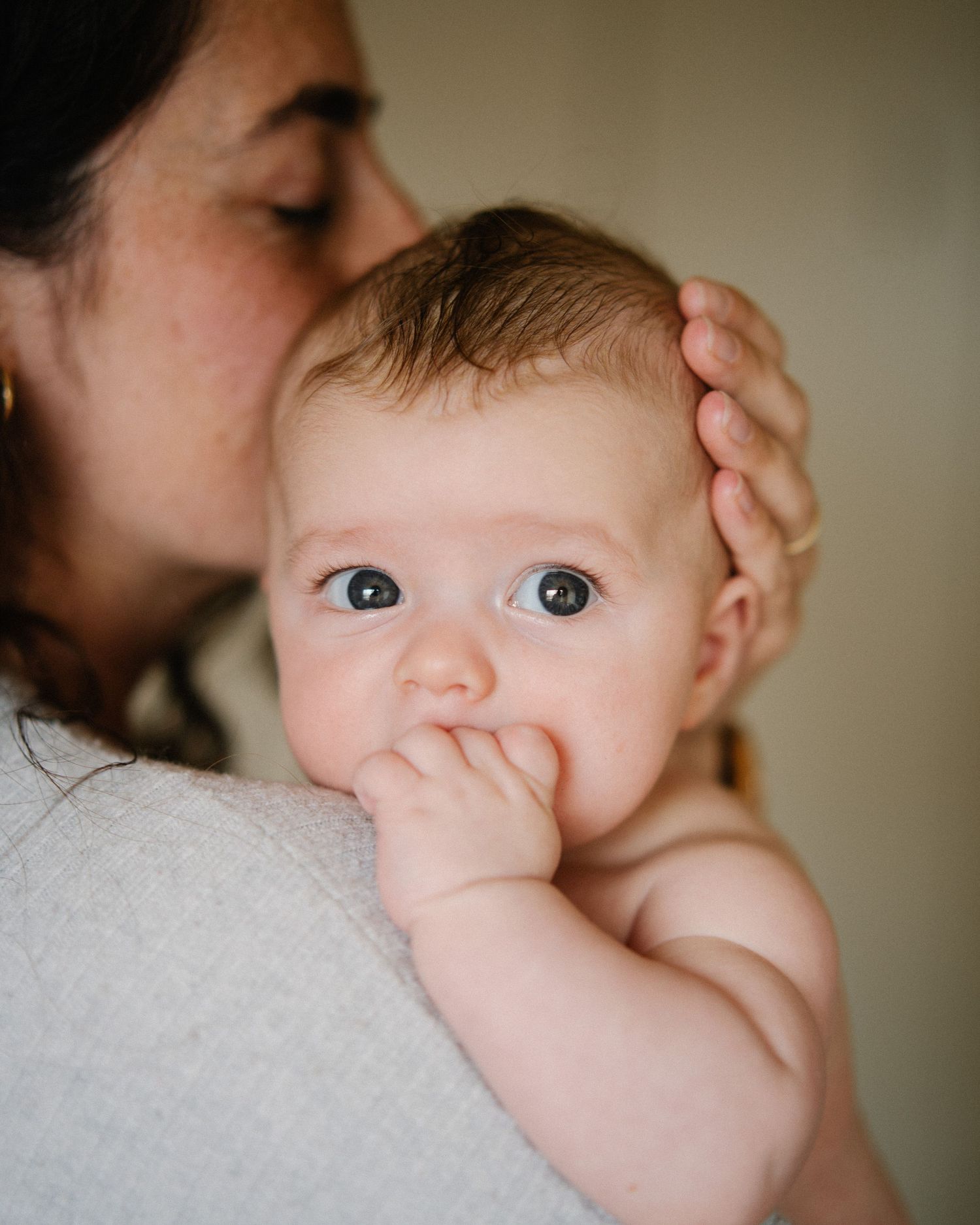 A tender parenting moment captured as a baby with bright eyes sucks their thumb while being held close.