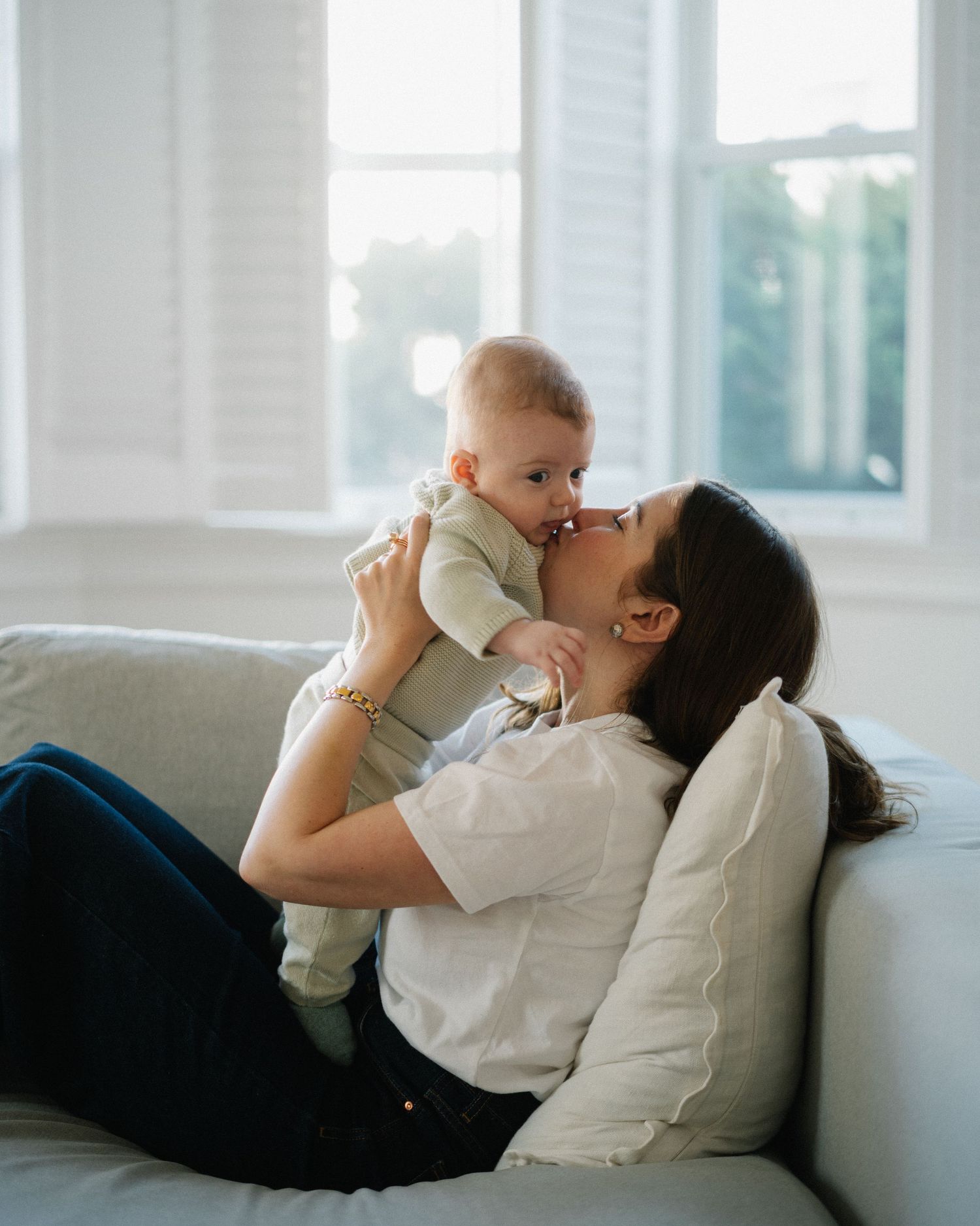 A mother lying on a white sofa lovingly kisses her baby while holding the infant up in a bright living room.