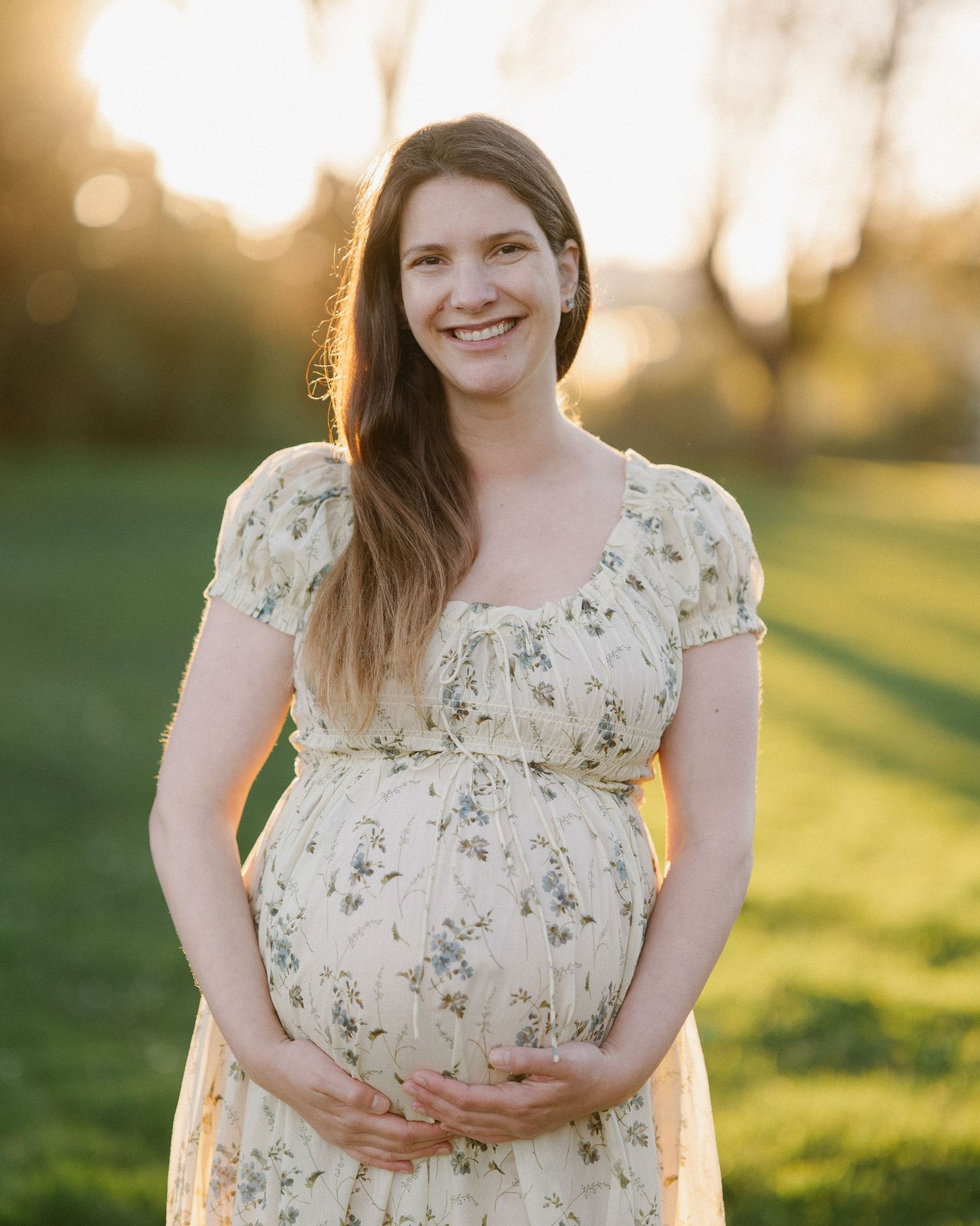 Woman in floral dress poses outdoors during golden hour sunset in a grassy field.