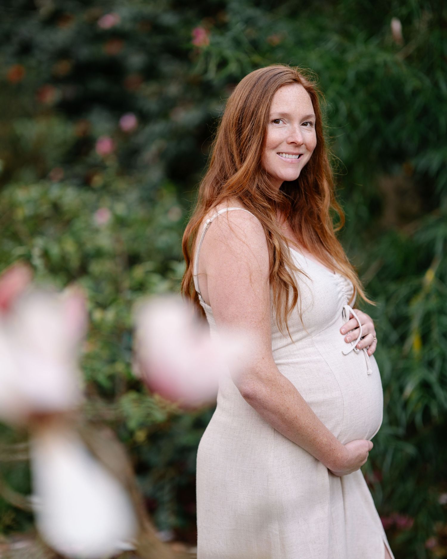 A person in a white dress poses outdoors against a natural green backdrop with blurred bokeh effects.