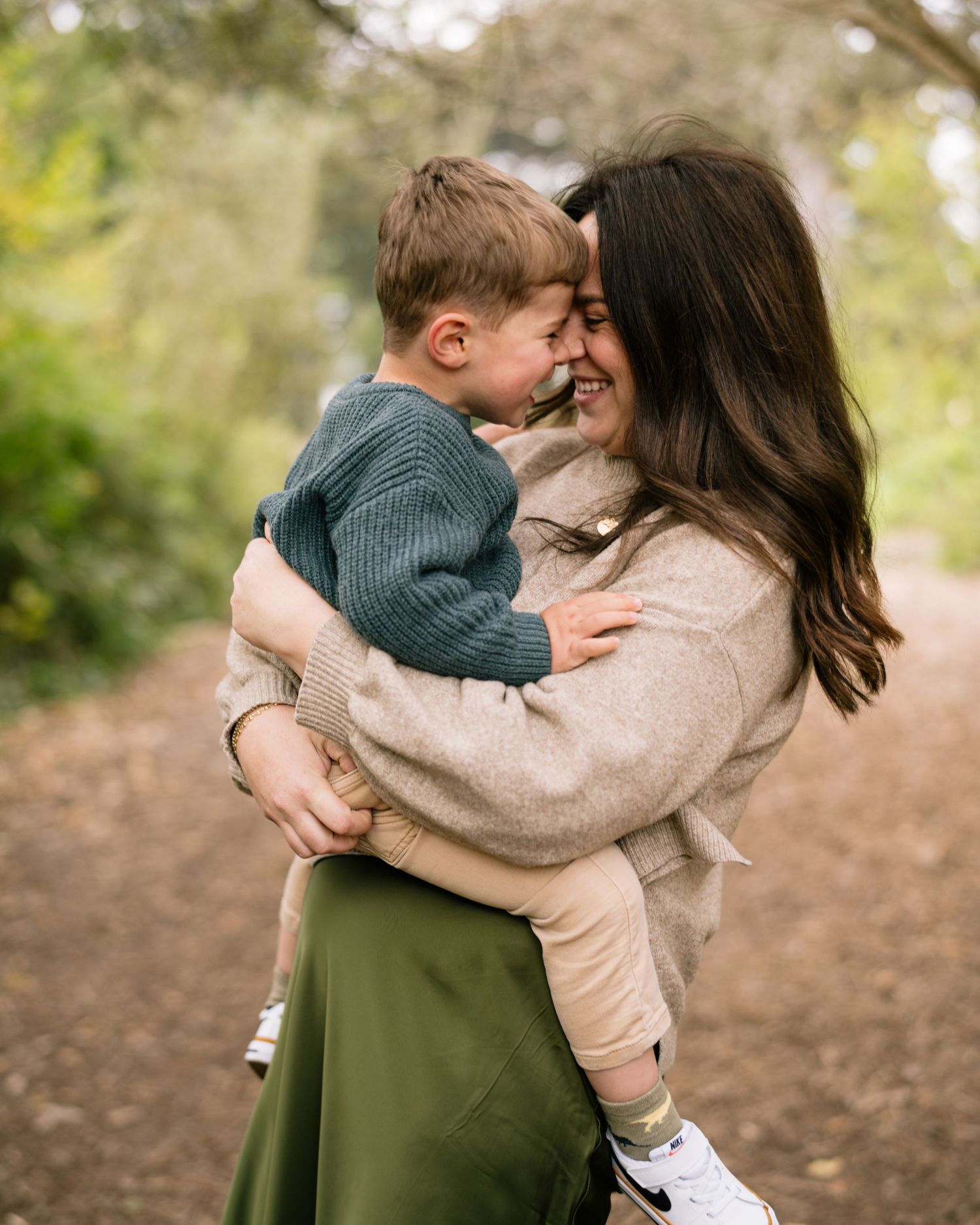 A smiling mother holds her laughing toddler boy on a nature path, both wearing cozy sweaters.