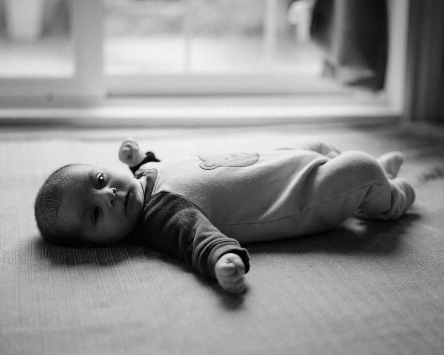 A baby in a light onesie lies on a hardwood floor in a black and white photograph.