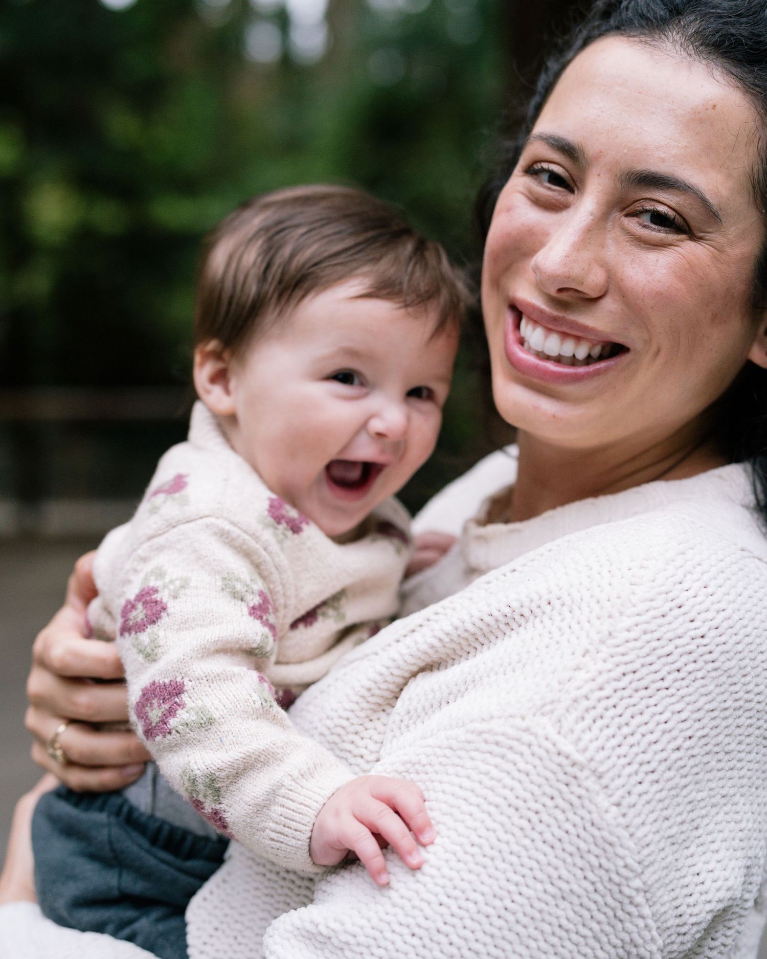 Smiling mother holding her laughing baby outdoors, both wearing cozy knit sweaters in a lush green setting.