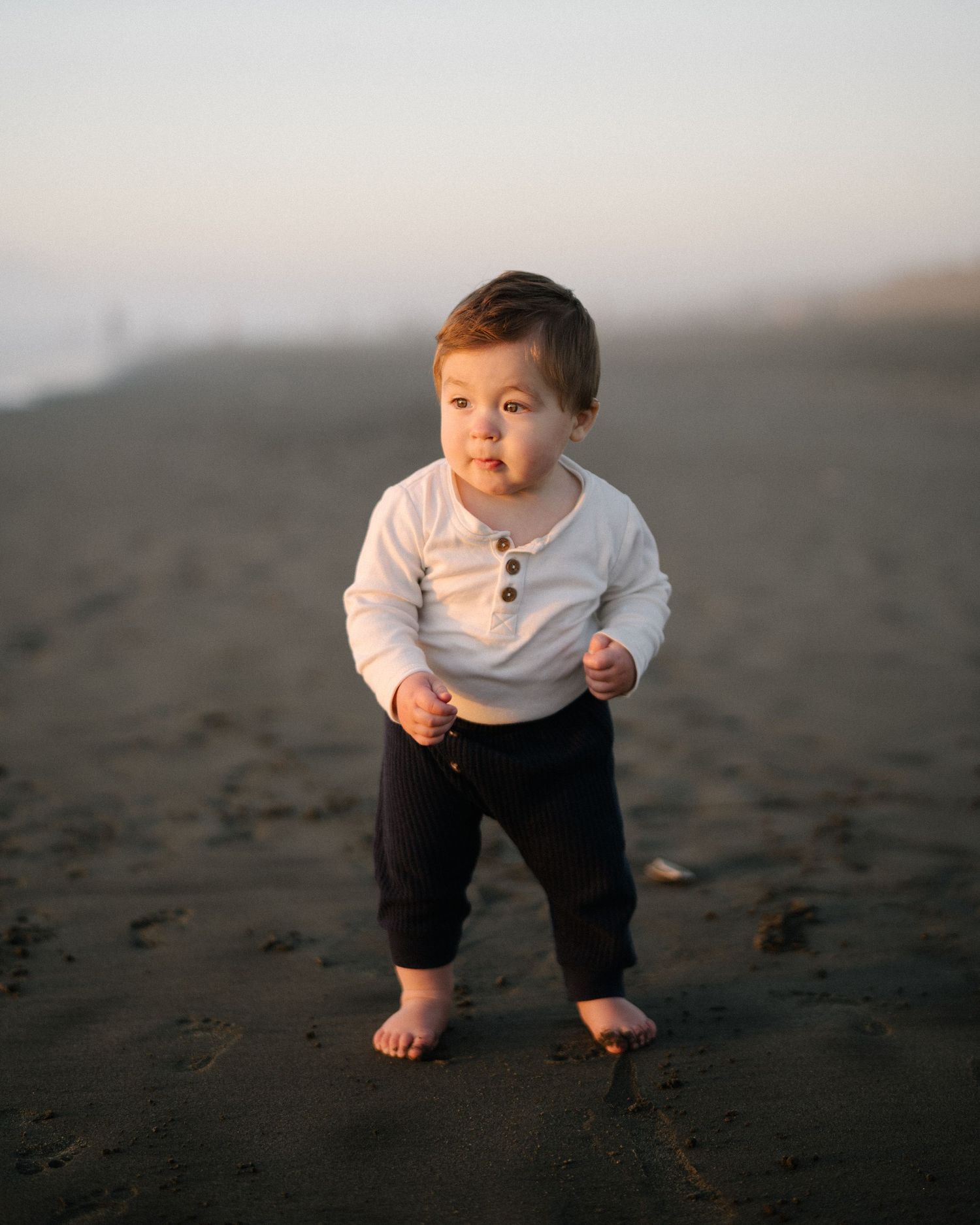 A small child in white clothing explores a dark sandy beach during golden hour.