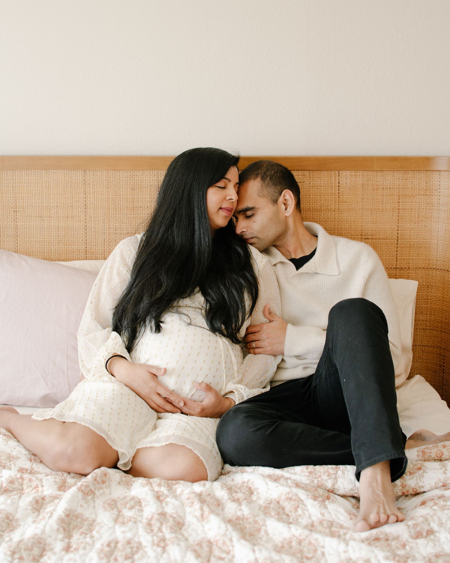 A couple shares a tender moment on a bed, with the woman cradling her pregnant belly as they lean close together.