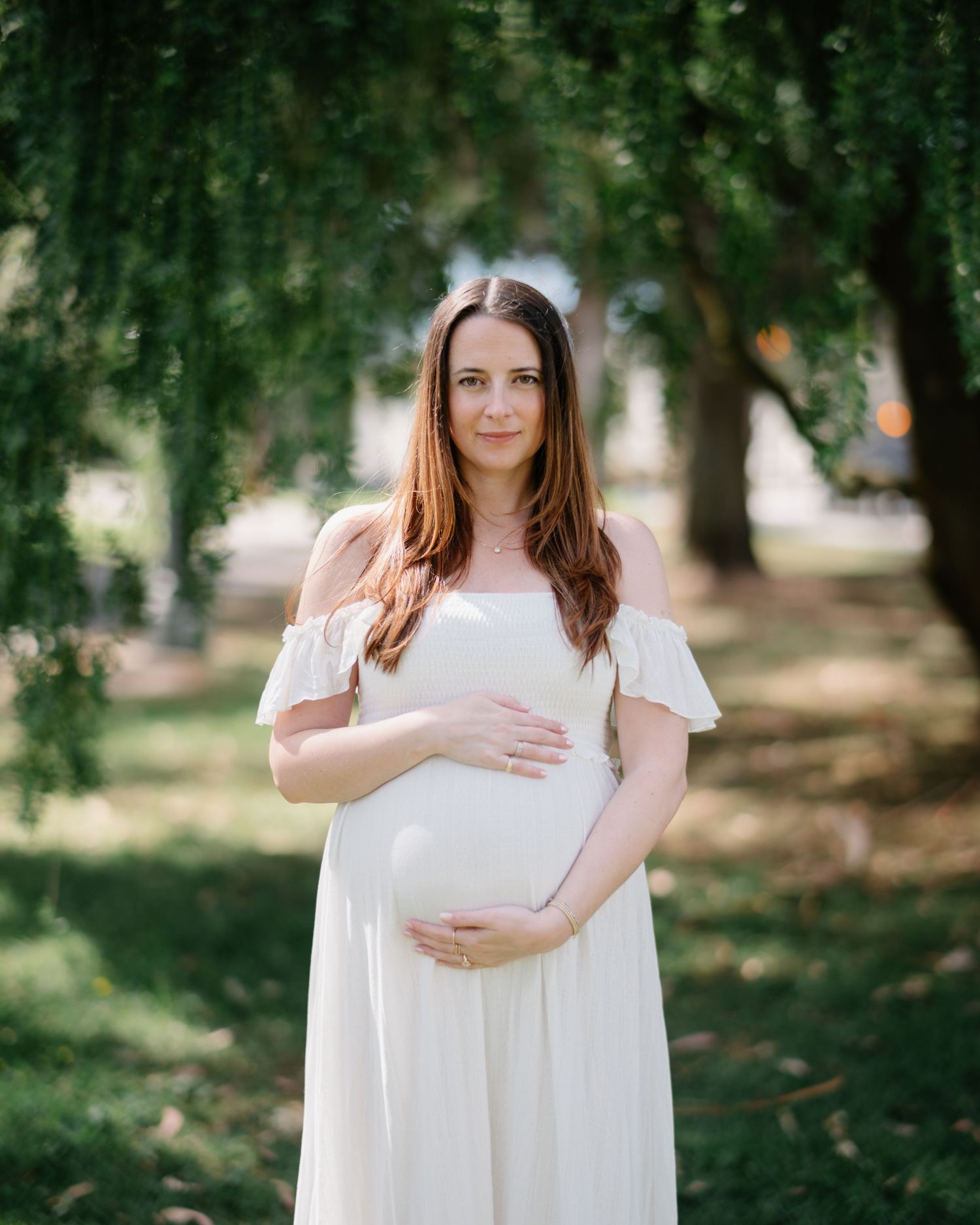 A pregnant woman in a flowing white dress stands serenely in a sunlit garden setting with green trees in the background.