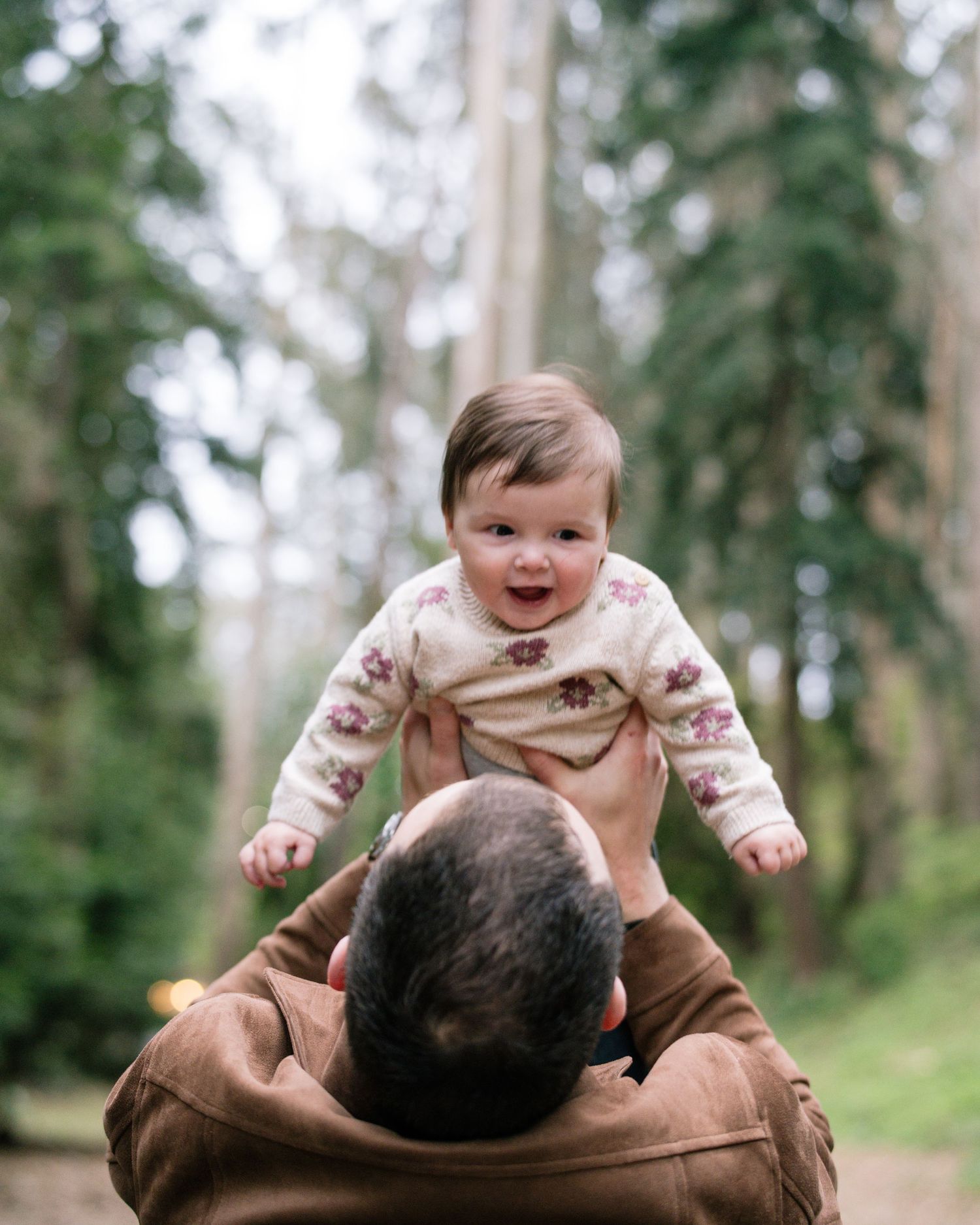Parent playfully lifts a laughing baby in the air against a backdrop of trees in a park setting.