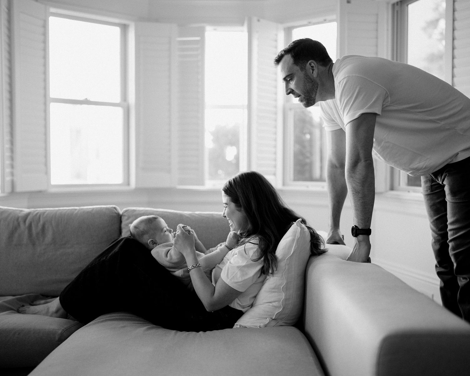 A tender black and white moment shared between a couple relaxing on a white couch by bright windows.