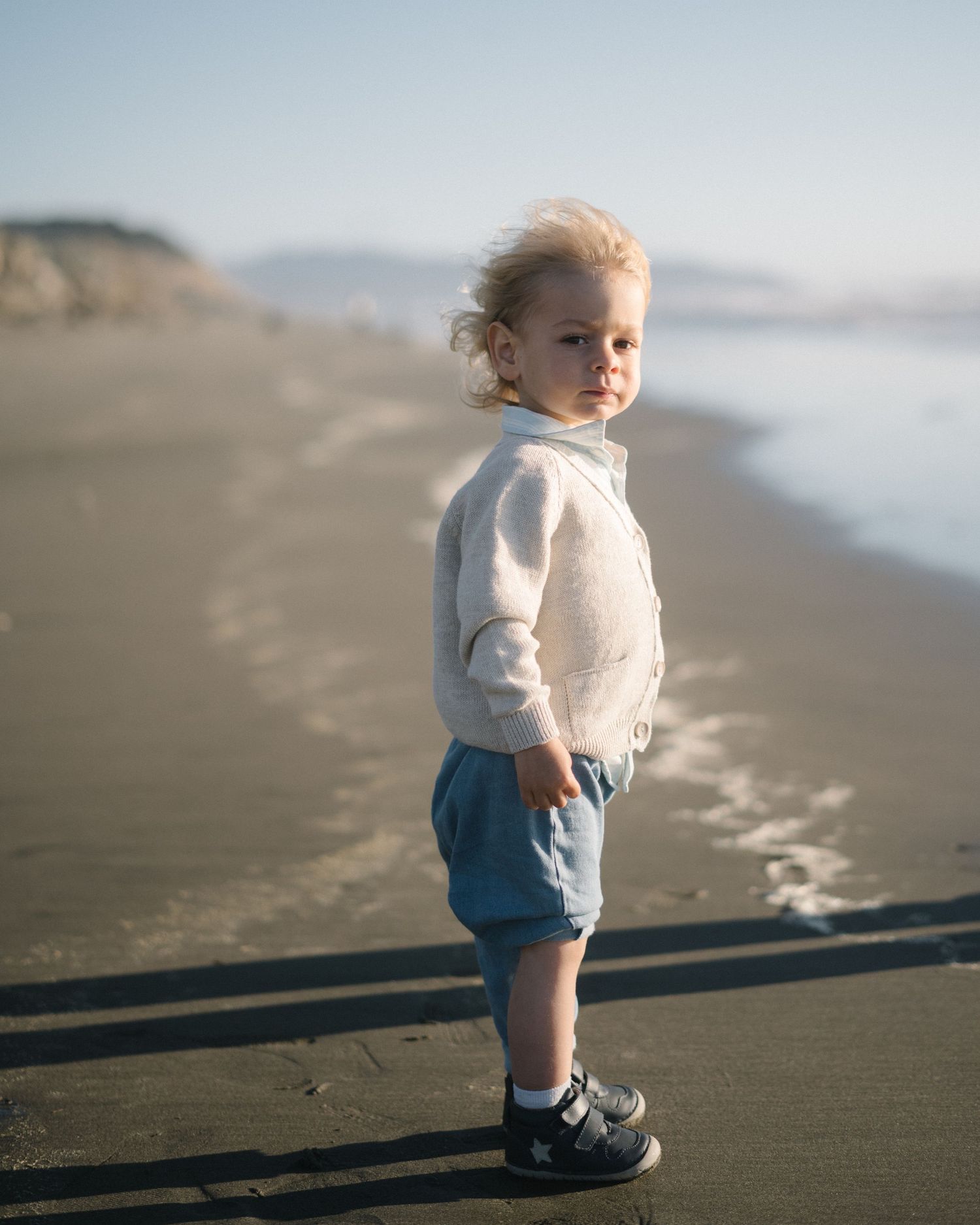 A small child in denim shorts and white top walks along a misty beach shoreline at sunset.