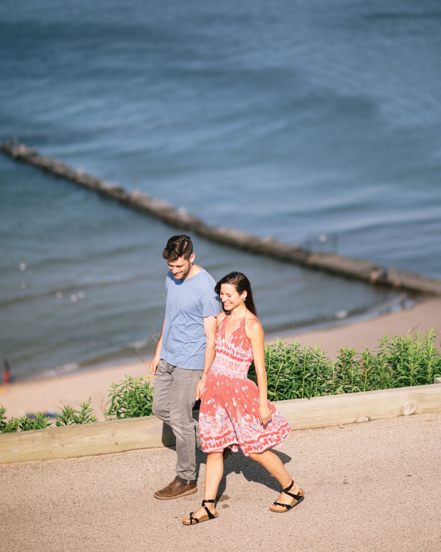 A couple walks along a beachside path with the ocean visible in the background on a sunny day.