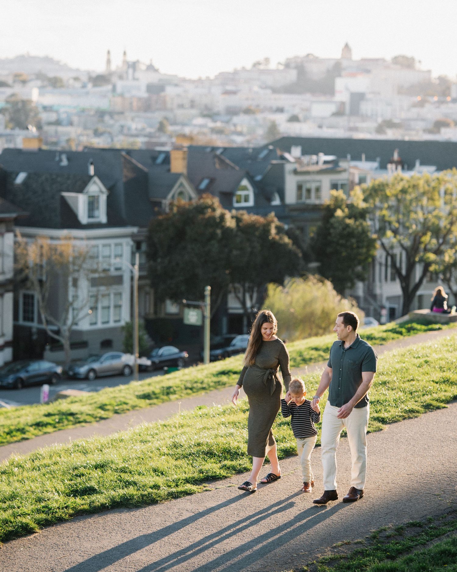 Family of three walking on a sunny grassy hill in San Francisco with Victorian homes and city skyline in the background.