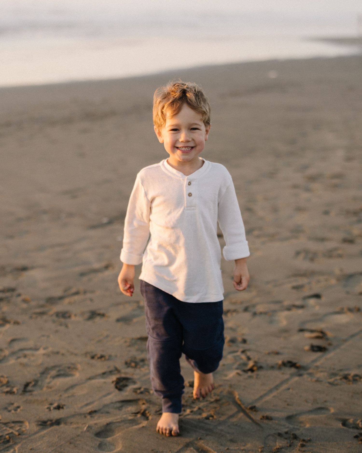 A young child in a white shirt and dark pants walks barefoot on a sandy beach at sunset.