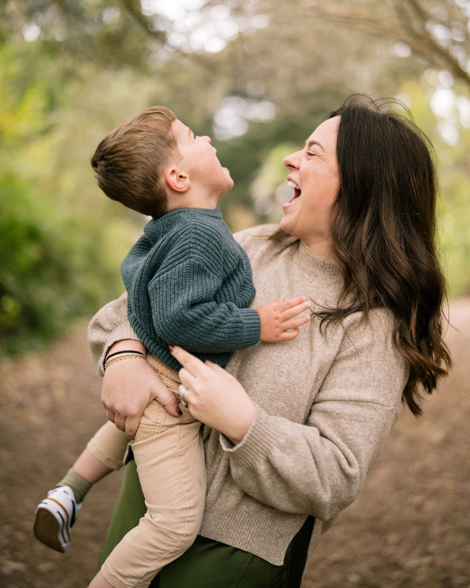 A joyful mother holds her laughing toddler boy outdoors on a wooded path, both sharing a playful moment.