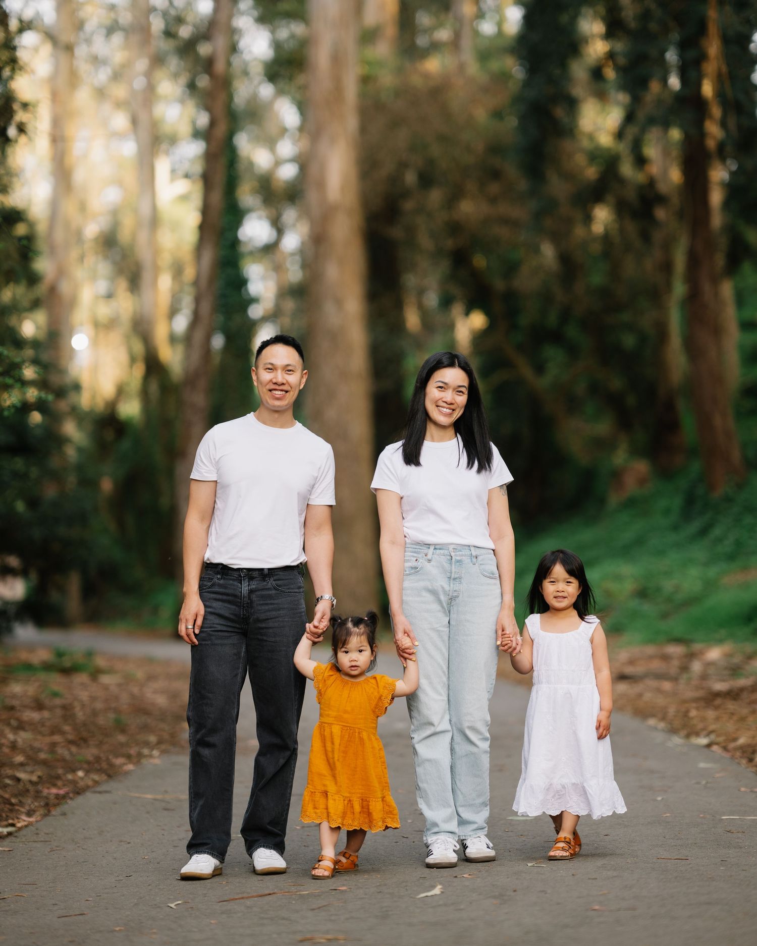 A smiling Asian family of four poses on a tree-lined path, parents in white tops with two young daughters in colorful dresses.