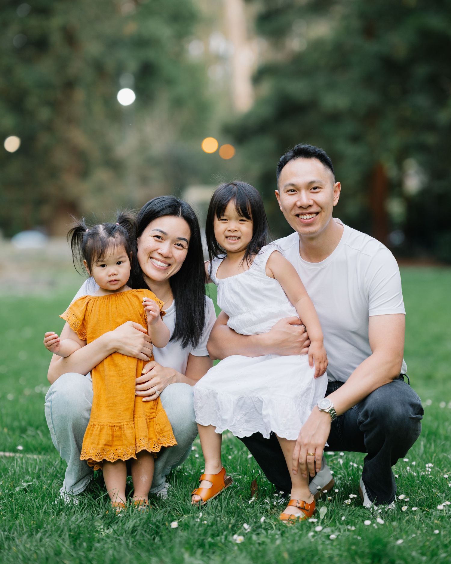 Happy Asian family of four poses outdoors on green grass, parents smiling with two young daughters in colorful dresses.