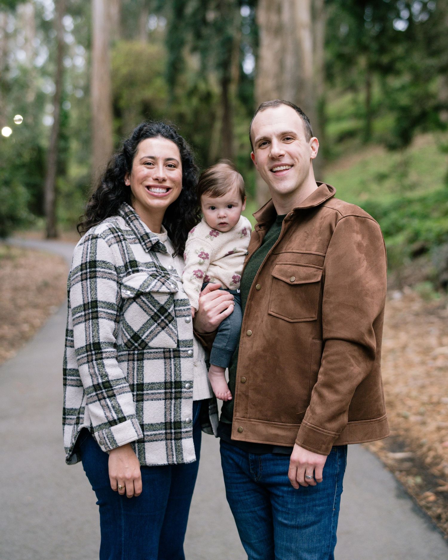 Family in matching plaid and brown outfits poses together on a tree-lined path during autumn.
