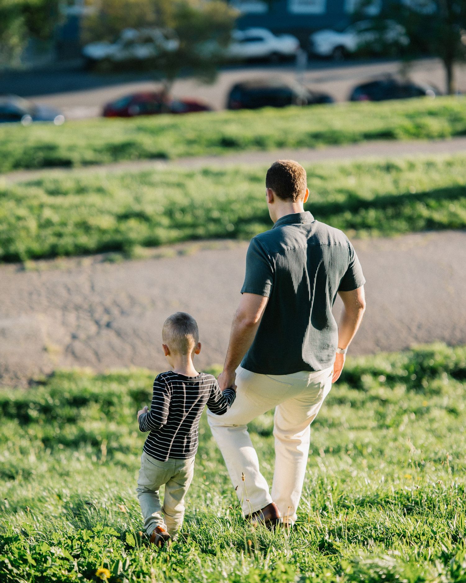 Adult man and young child walking hand in hand across a green grassy park on a sunny day.