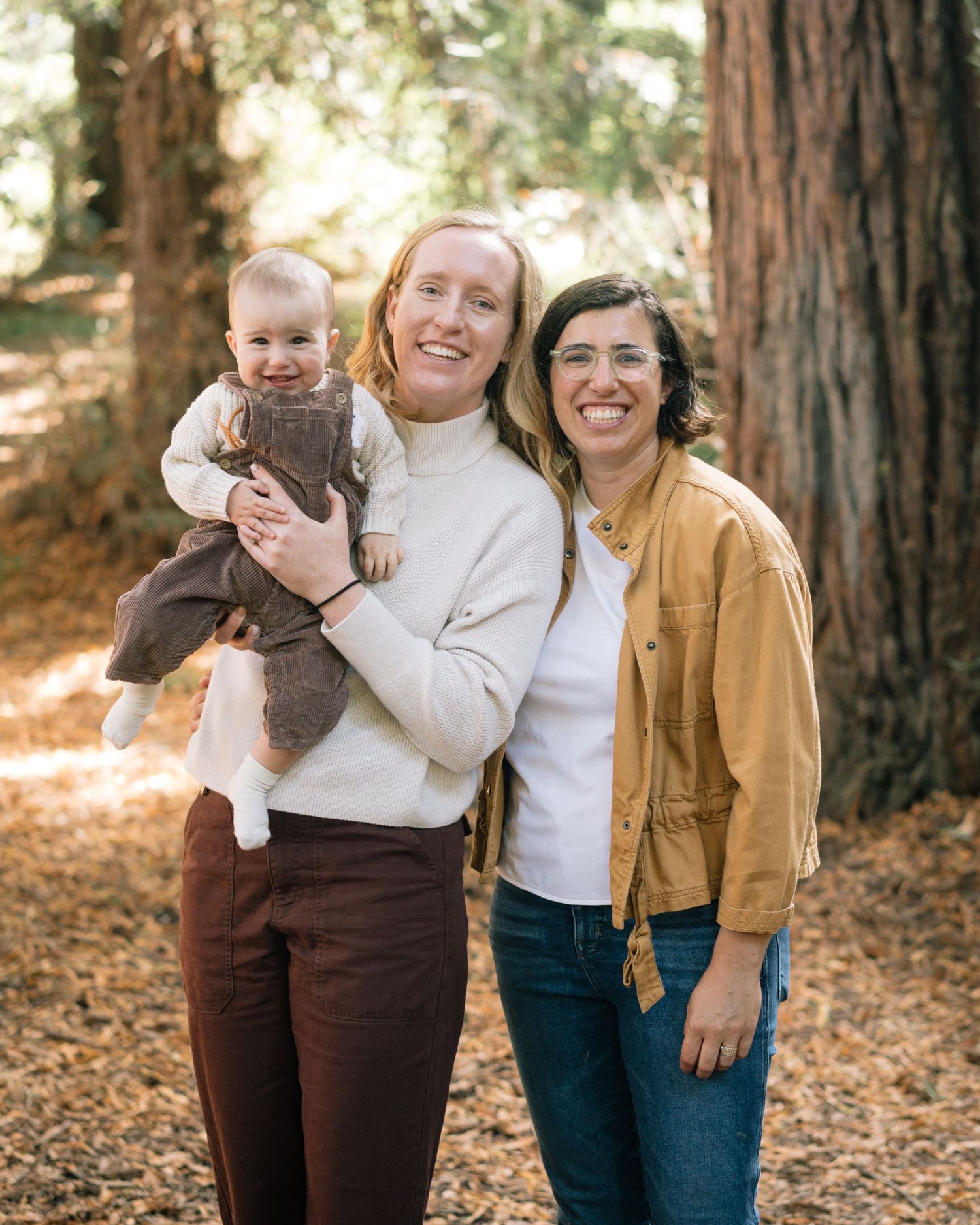 Three people stand together on fallen autumn leaves in a forest of tall redwood trees.