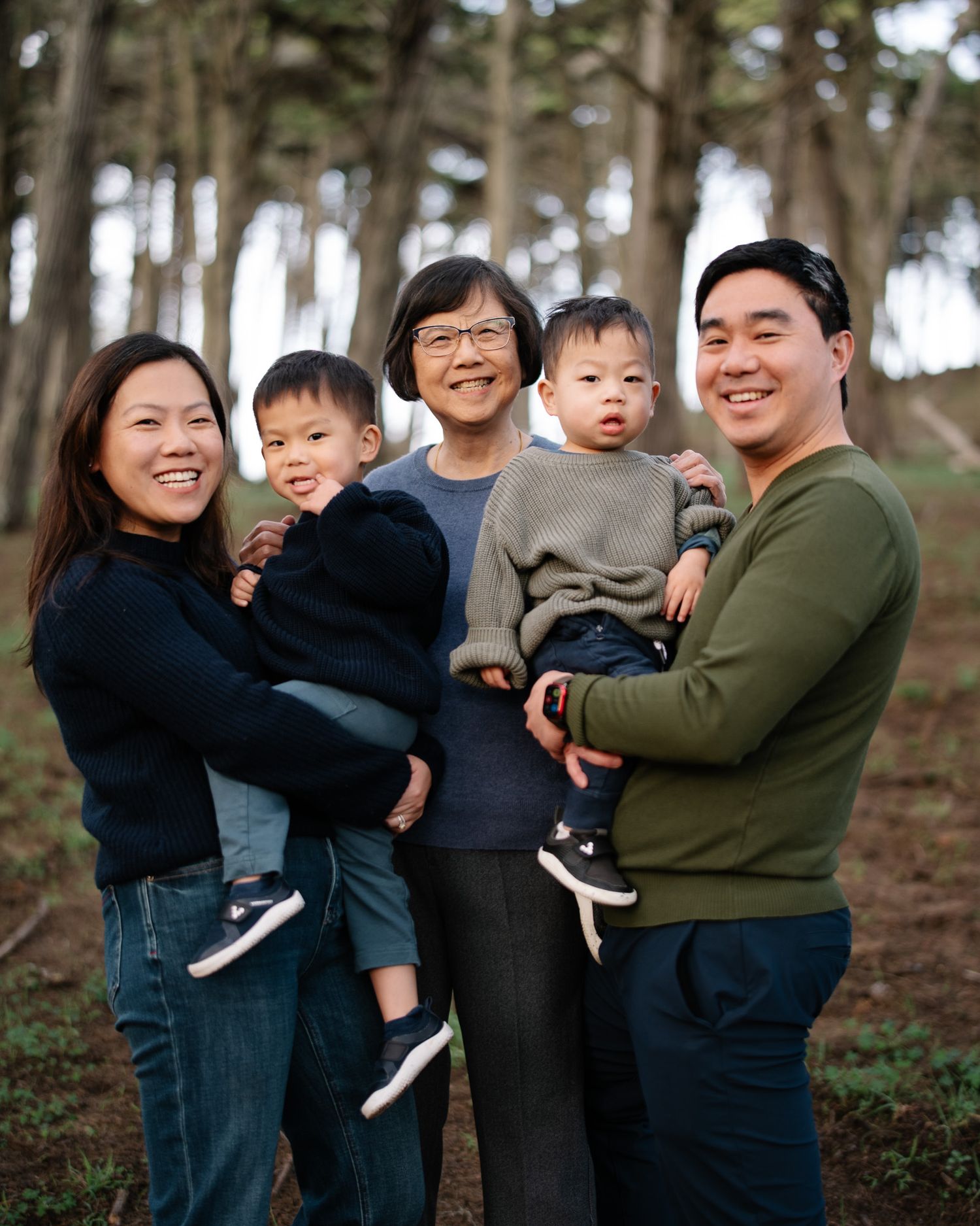 A joyful family photoshoot in a wooded park setting shows candid moments of laughter and embraces.