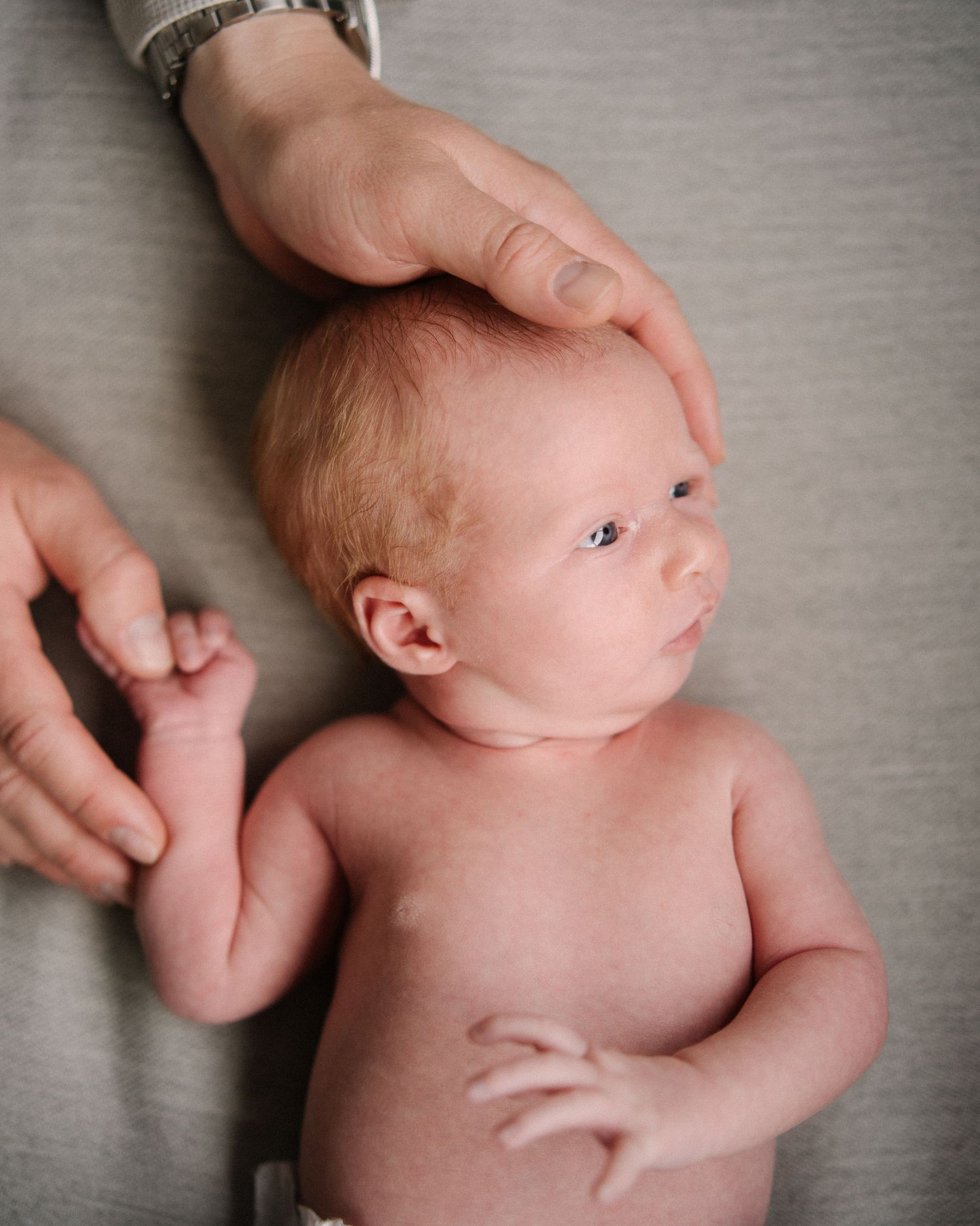 A gentle hand touches the head of a newborn baby lying on soft gray fabric.