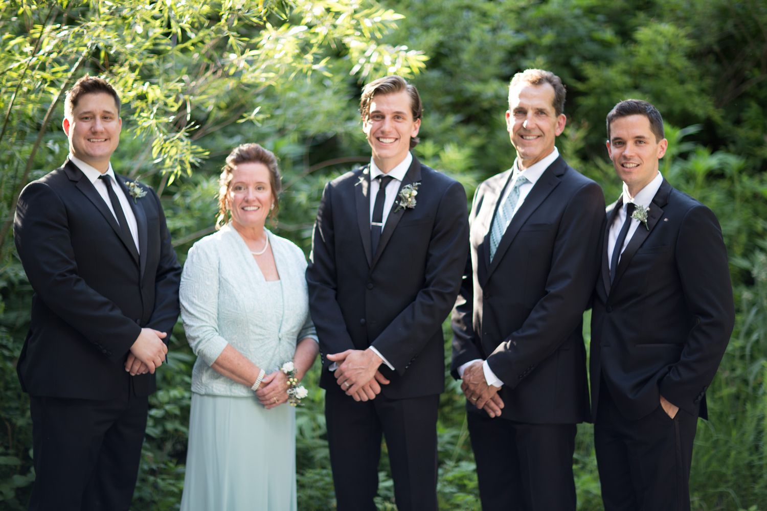 A wedding party in formal attire poses together outdoors against a lush green garden backdrop.