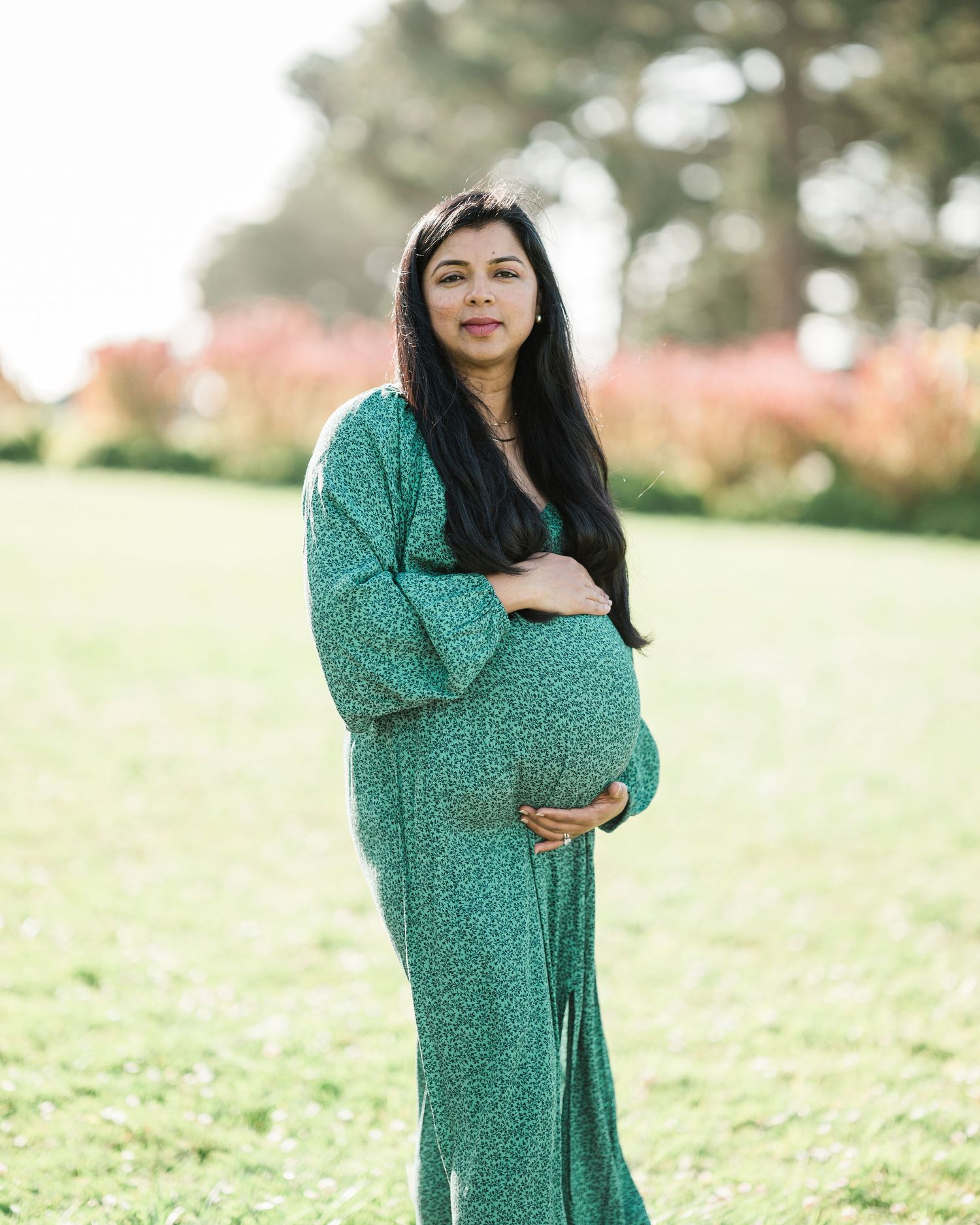 Pregnant woman in green floral maxi dress cradling baby bump outdoors in a lush garden setting.