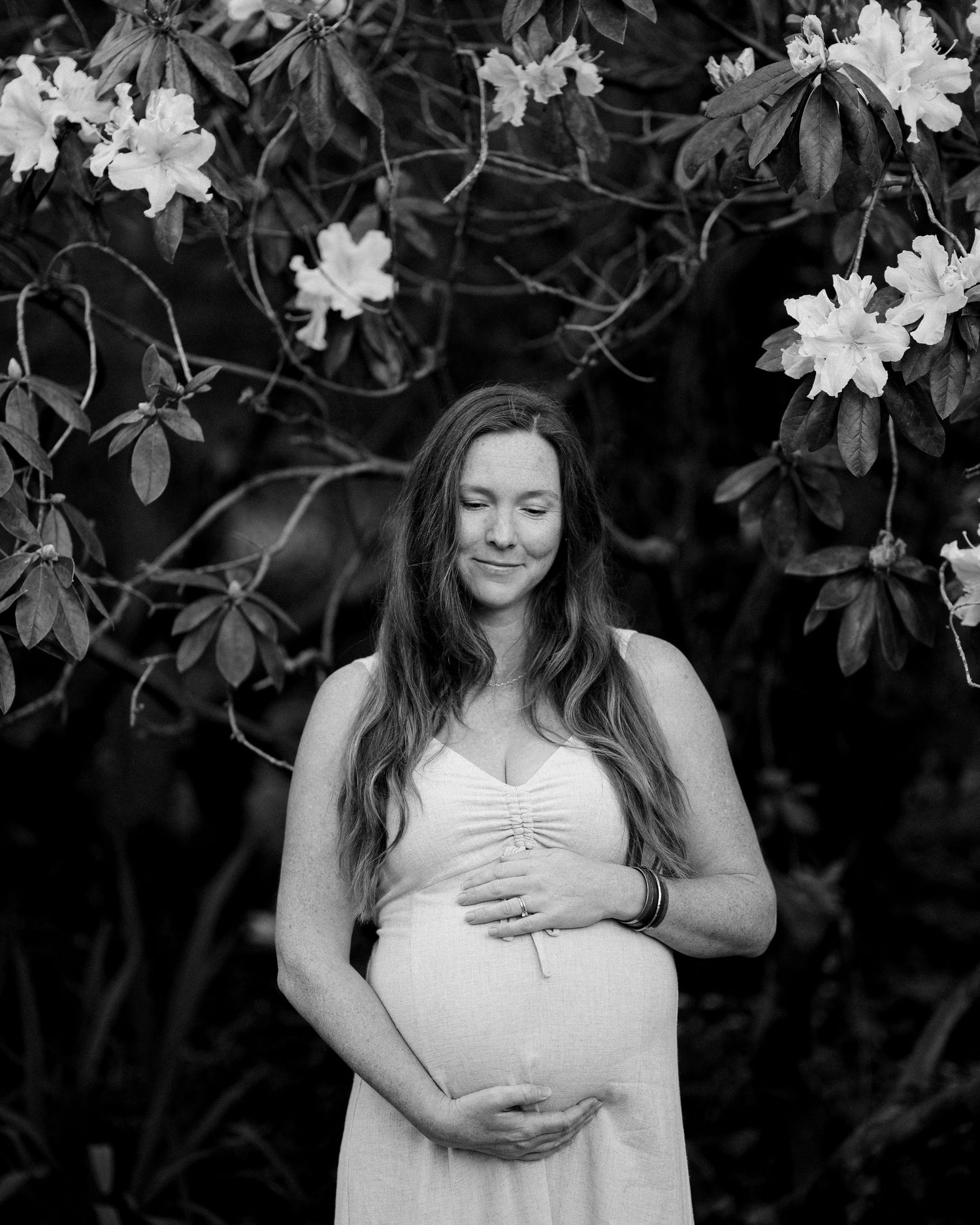 Black and white maternity photo series showing a woman in a white dress posing against a leafy backdrop.