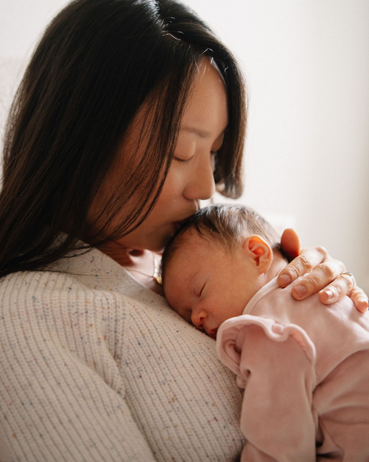 A mother tenderly kisses her sleeping newborn baby dressed in pink, sharing a peaceful bonding moment.