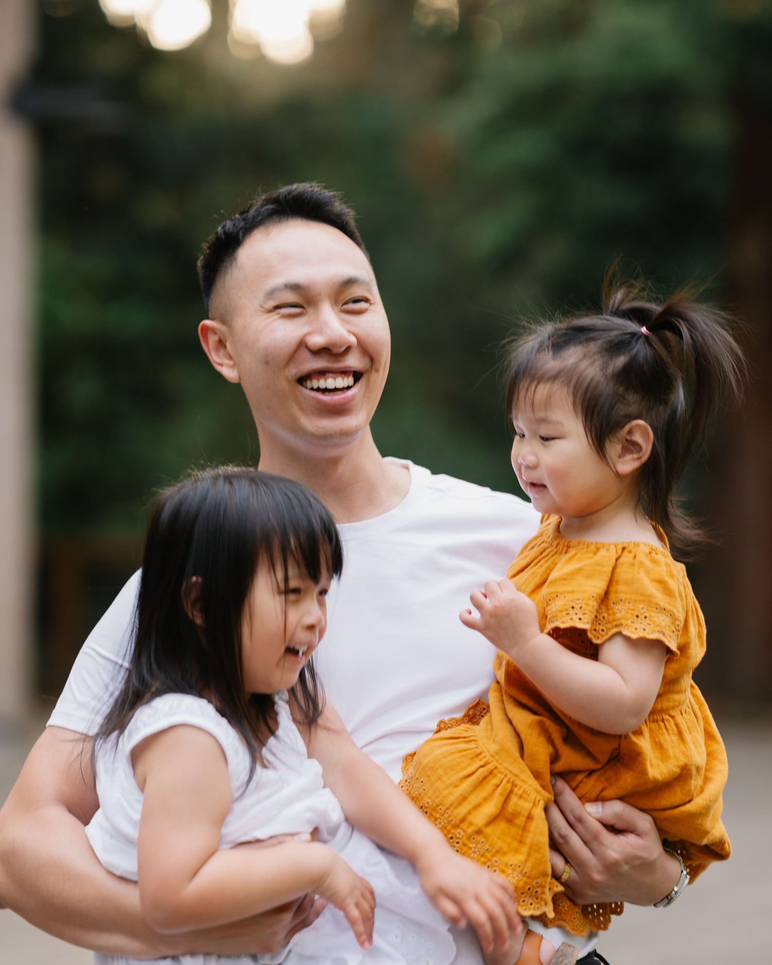 A smiling Asian man in a white t-shirt holds two young girls, one in white and one in an orange dress, outdoors.
