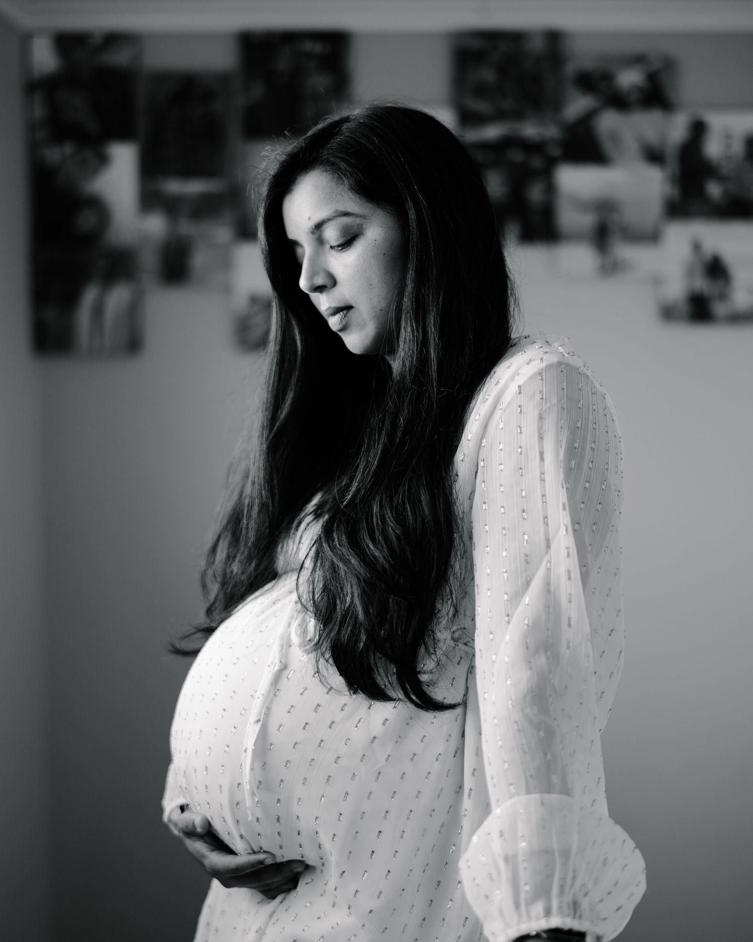 Black and white portrait of a pregnant woman in white dress cradling her baby bump, looking down thoughtfully.