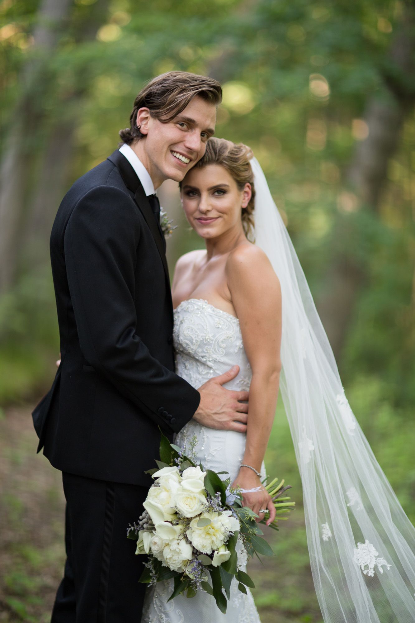 A bride and groom embrace in a sunlit wooded area with white floral bouquet.