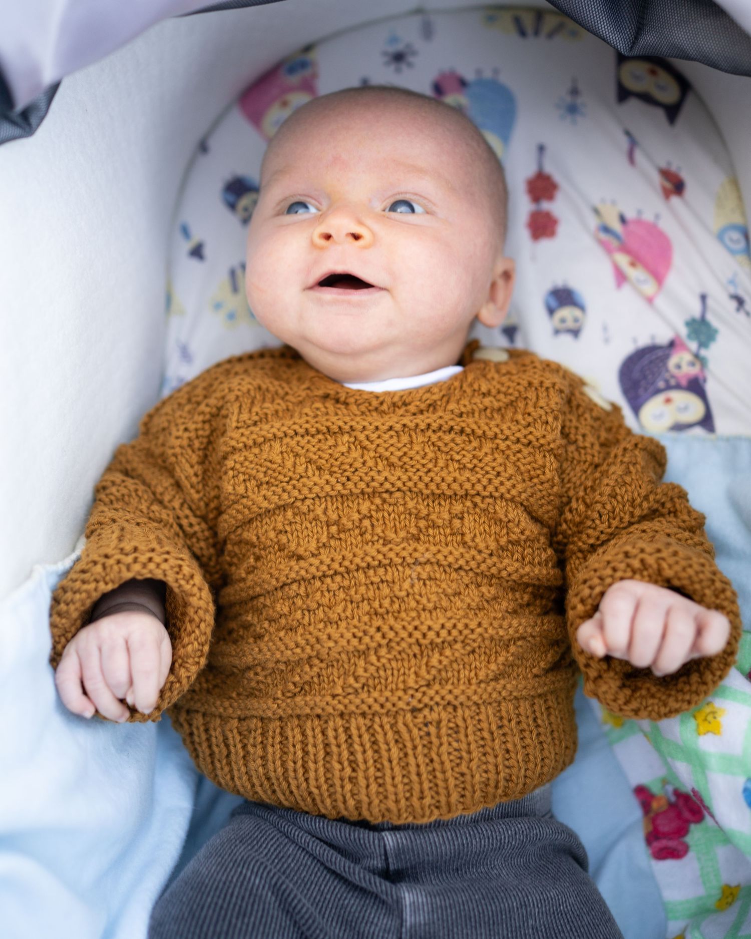 Baby wearing cozy brown knitted sweater and gray pants lying on floral patterned bedding.