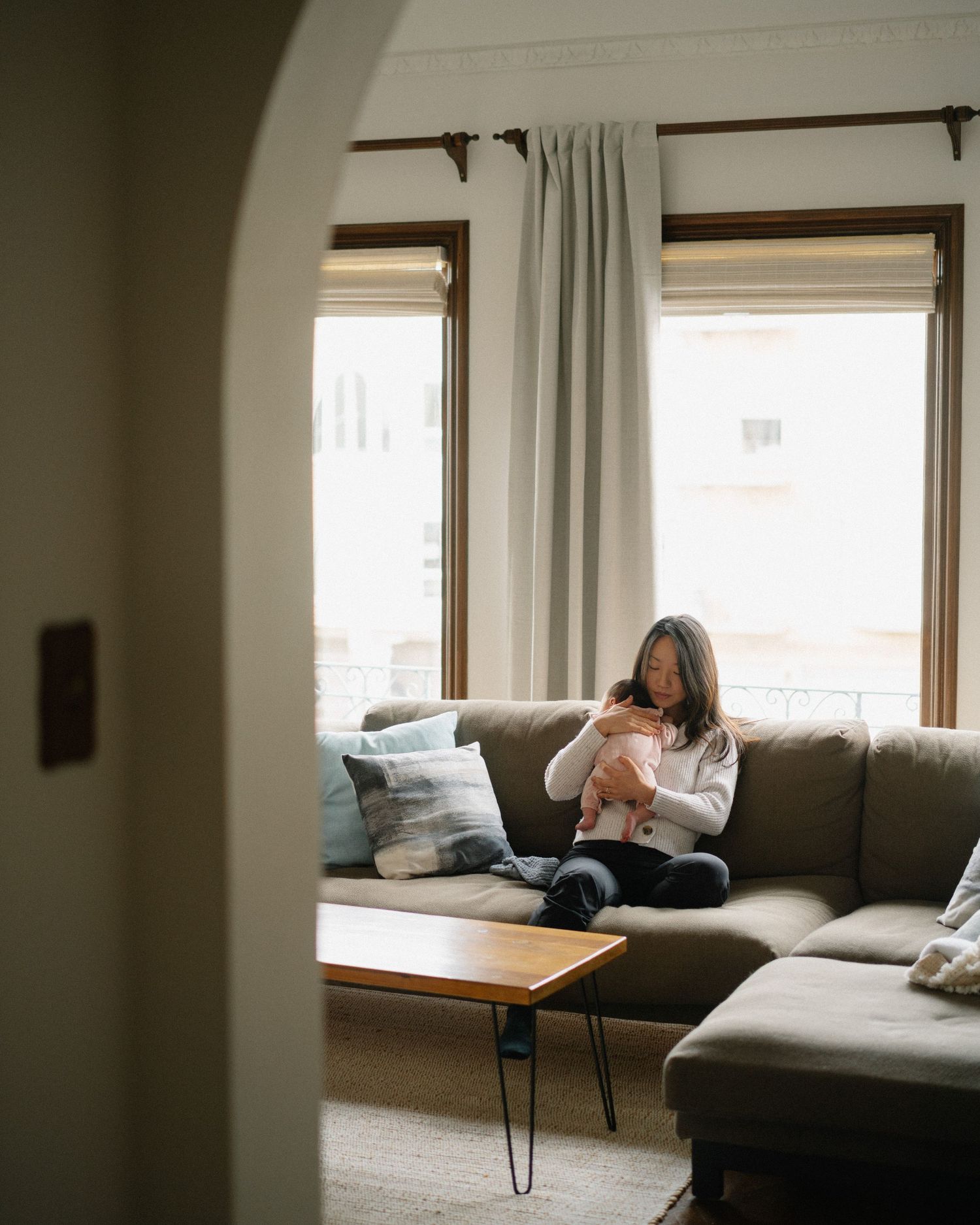 Woman sitting on a gray sectional sofa holding a baby in a cozy living room with large windows.