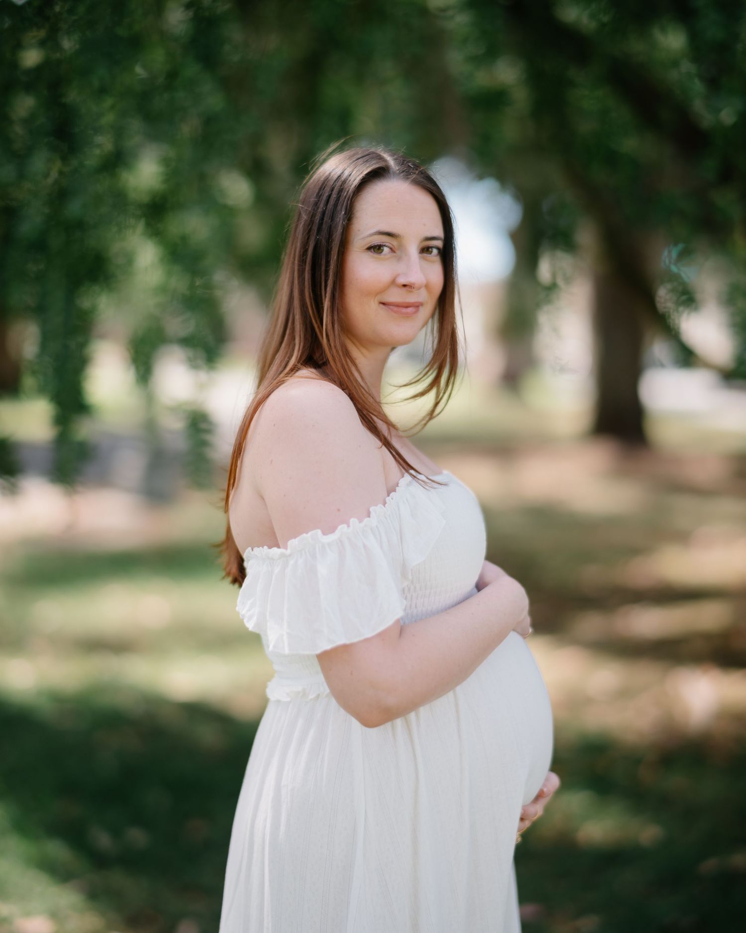 A maternity photoshoot in a white off-shoulder dress against a natural outdoor backdrop with trees and sunlight.