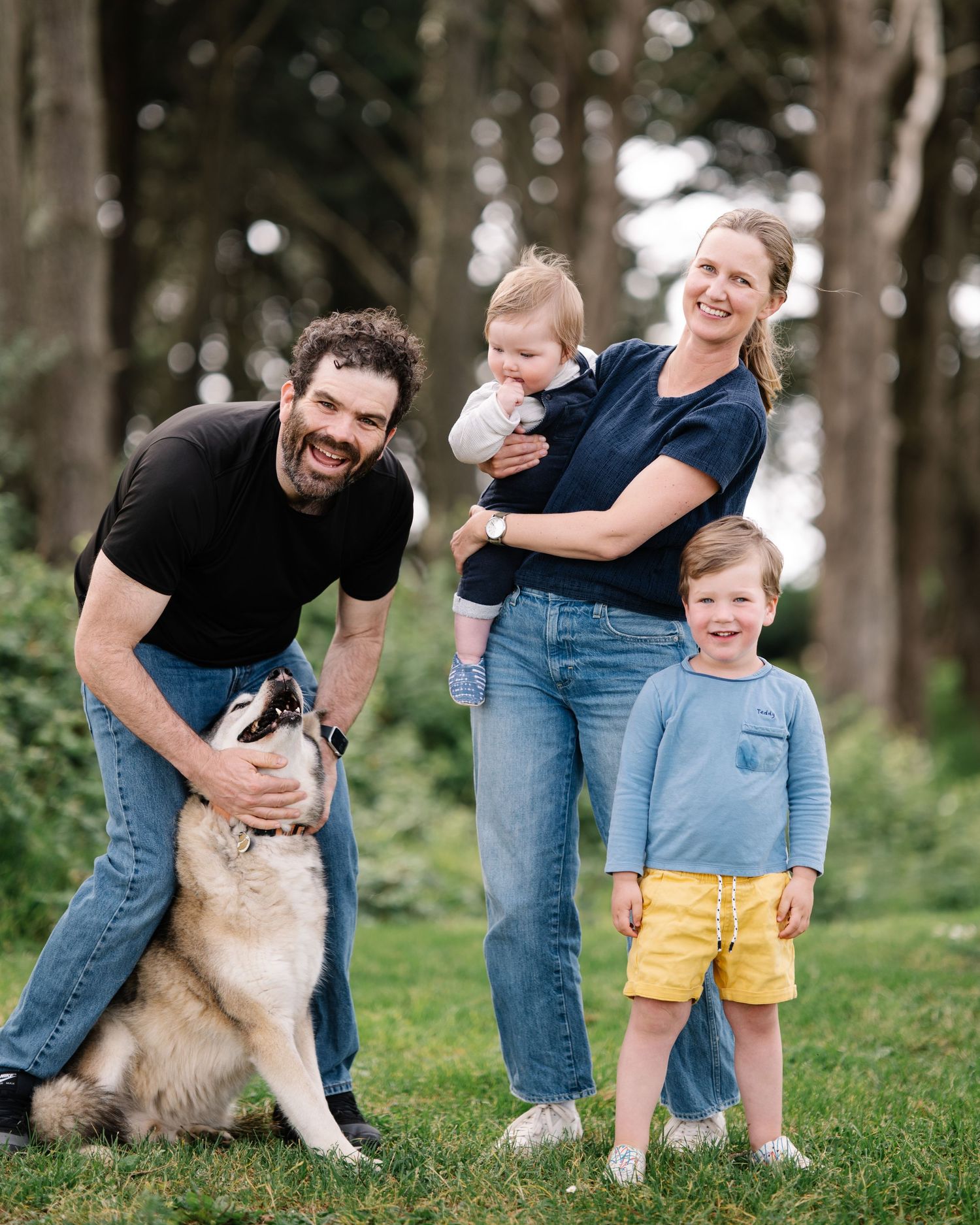 Happy family with two young children and a dog posing outdoors in a lush green park surrounded by trees.