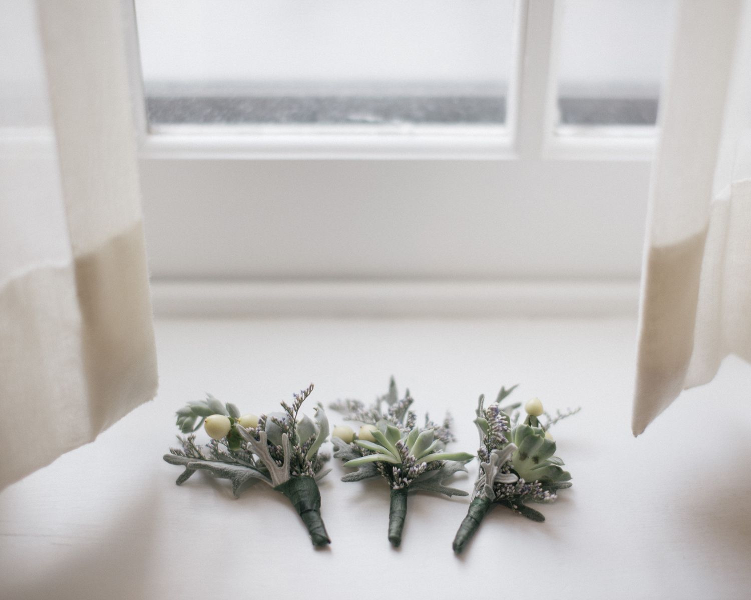Wedding boutonnieres with greenery arranged on a white windowsill.
