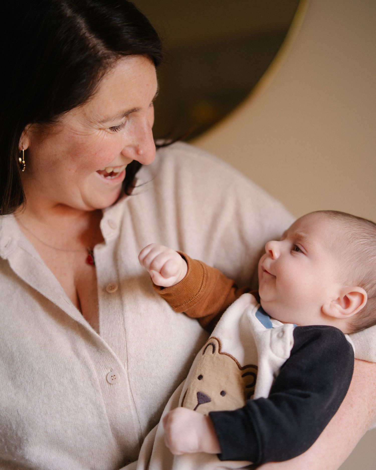 A woman in a beige cardigan smiles lovingly at a happy baby wearing a bear onesie cradled in her arms.
