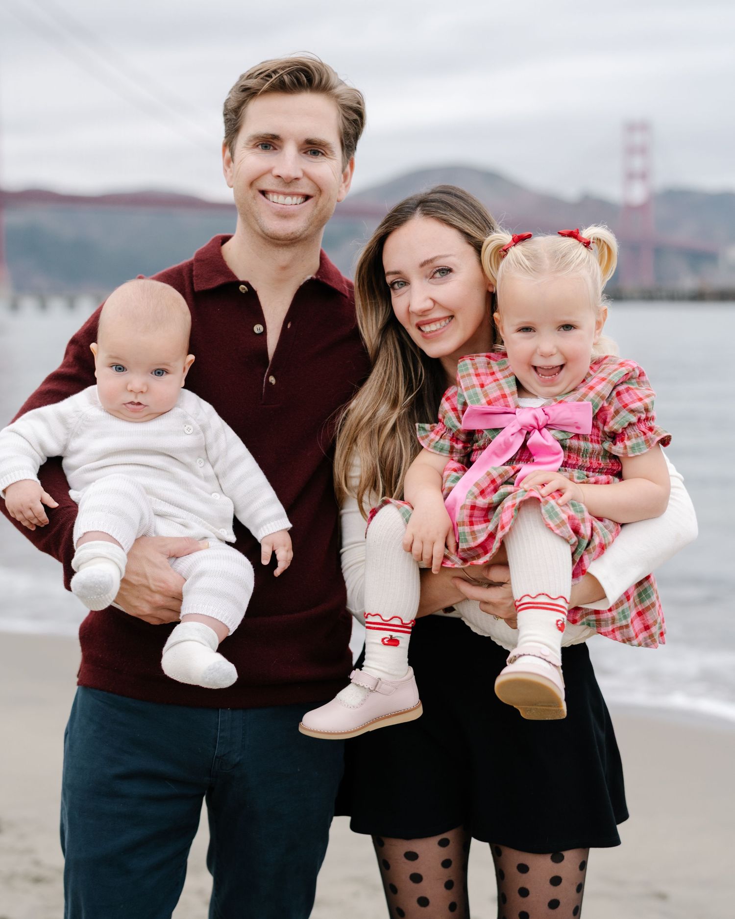 Family poses together at beach with Golden Gate Bridge visible in background on cloudy San Francisco day.