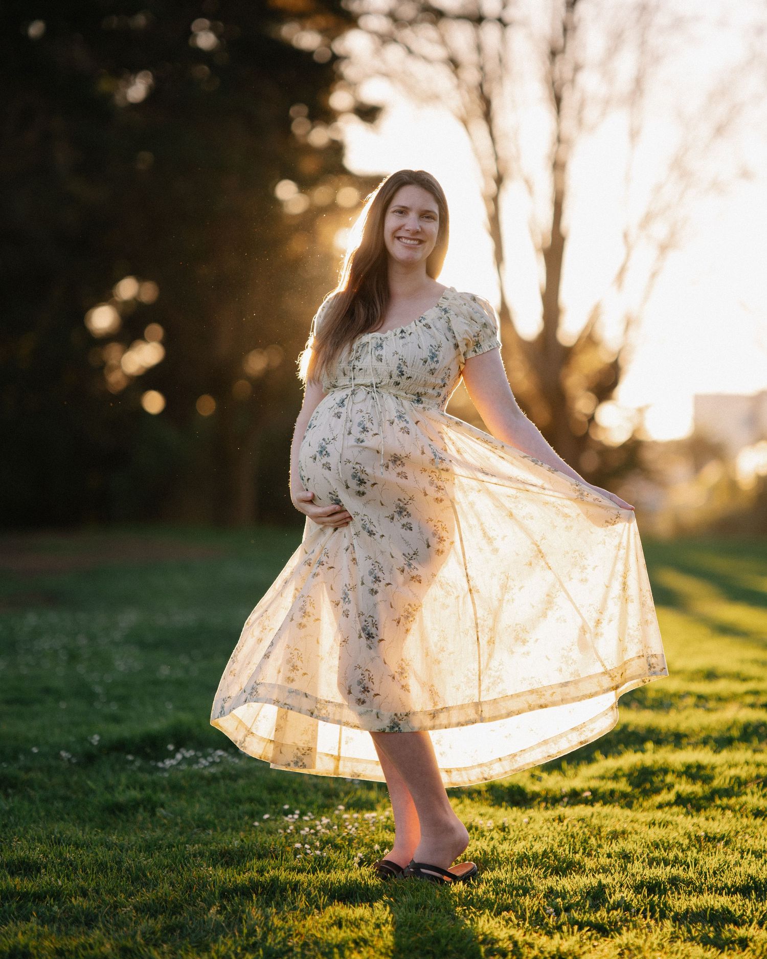 The hem of a flowing floral dress twirls against green grass in warm evening light.