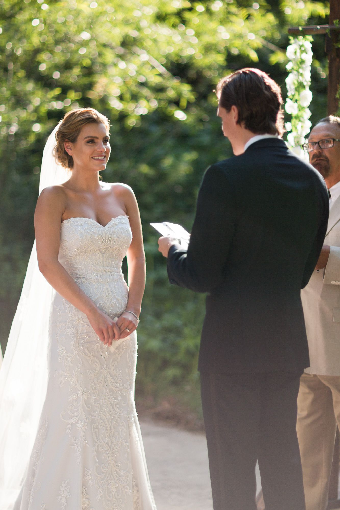 A wedding ceremony takes place outdoors with a bride in a white lace gown exchanging vows with the groom.