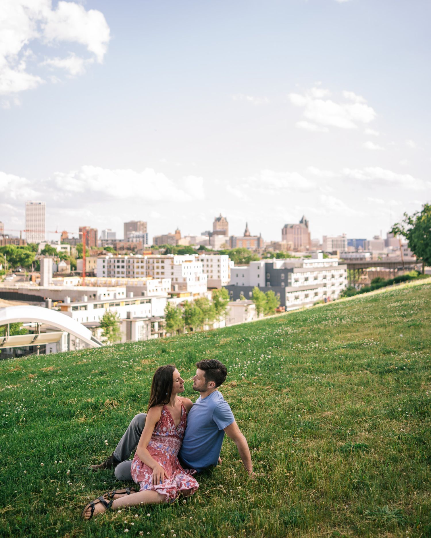 Two people sit on a grassy hillside overlooking a city skyline on a sunny summer afternoon.