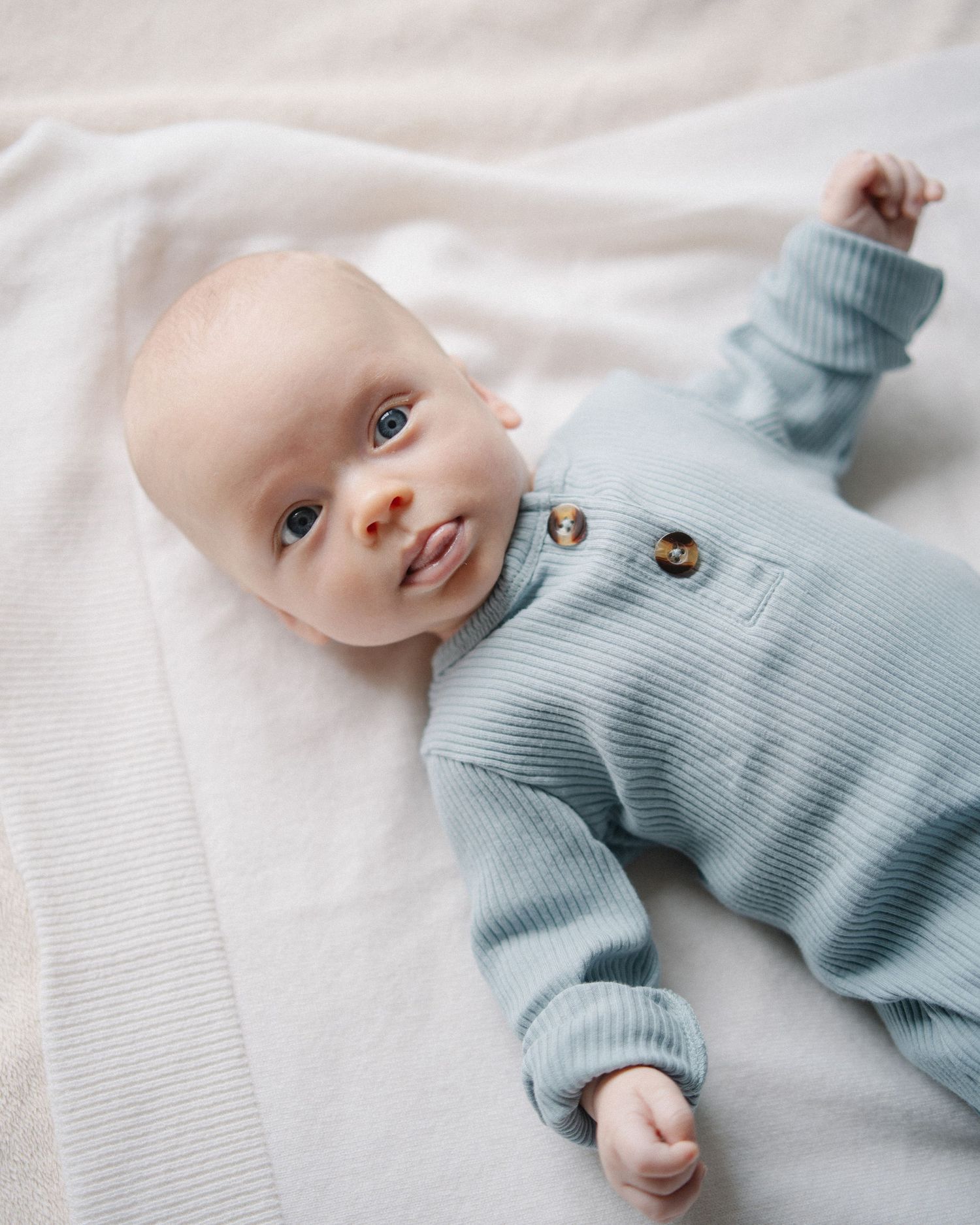 Baby wearing soft blue ribbed sweater lying on white textured fabric looking upward with bright eyes.