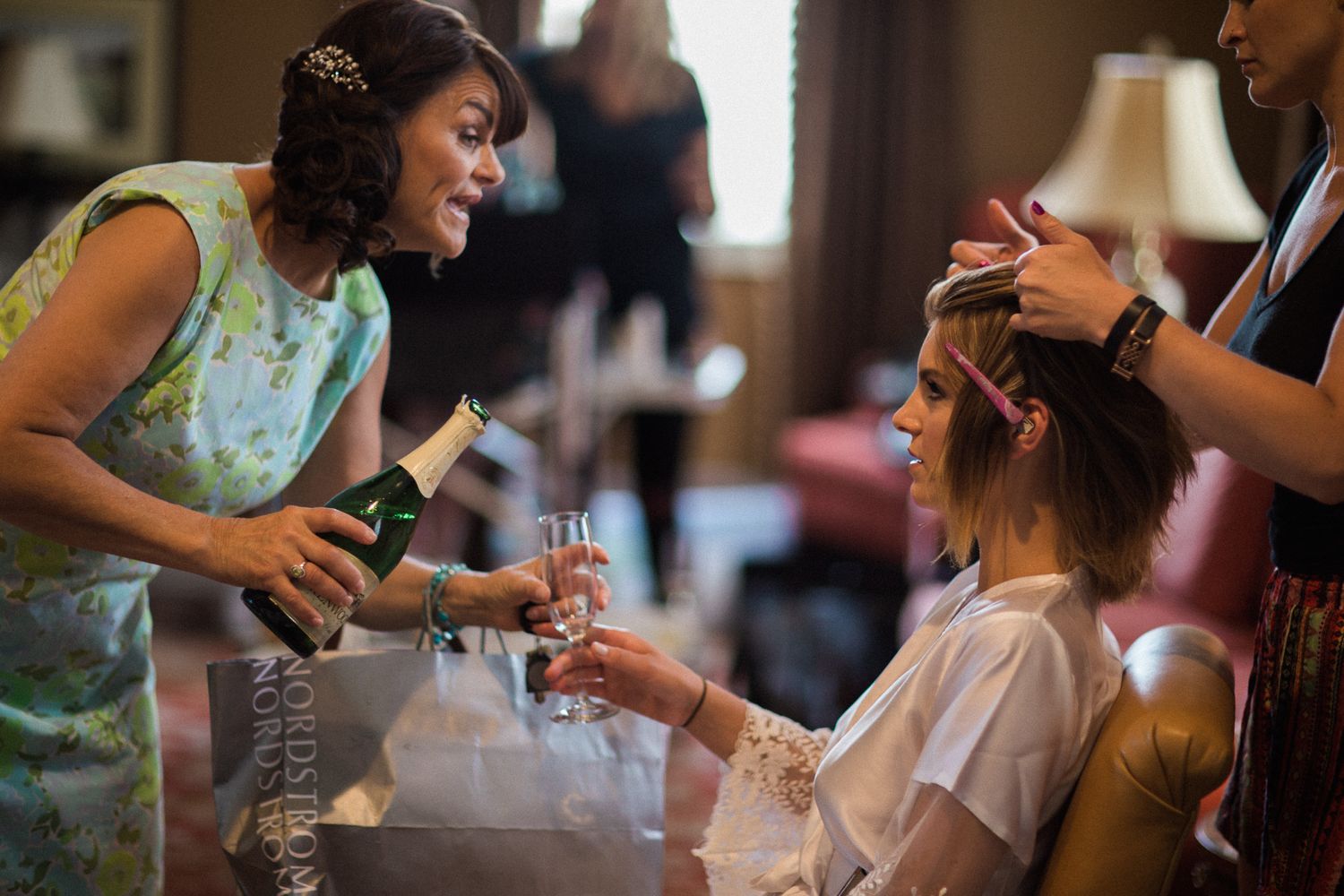 A hairstylist works on styling while someone holds a bottle and glass during preparation for a special event.