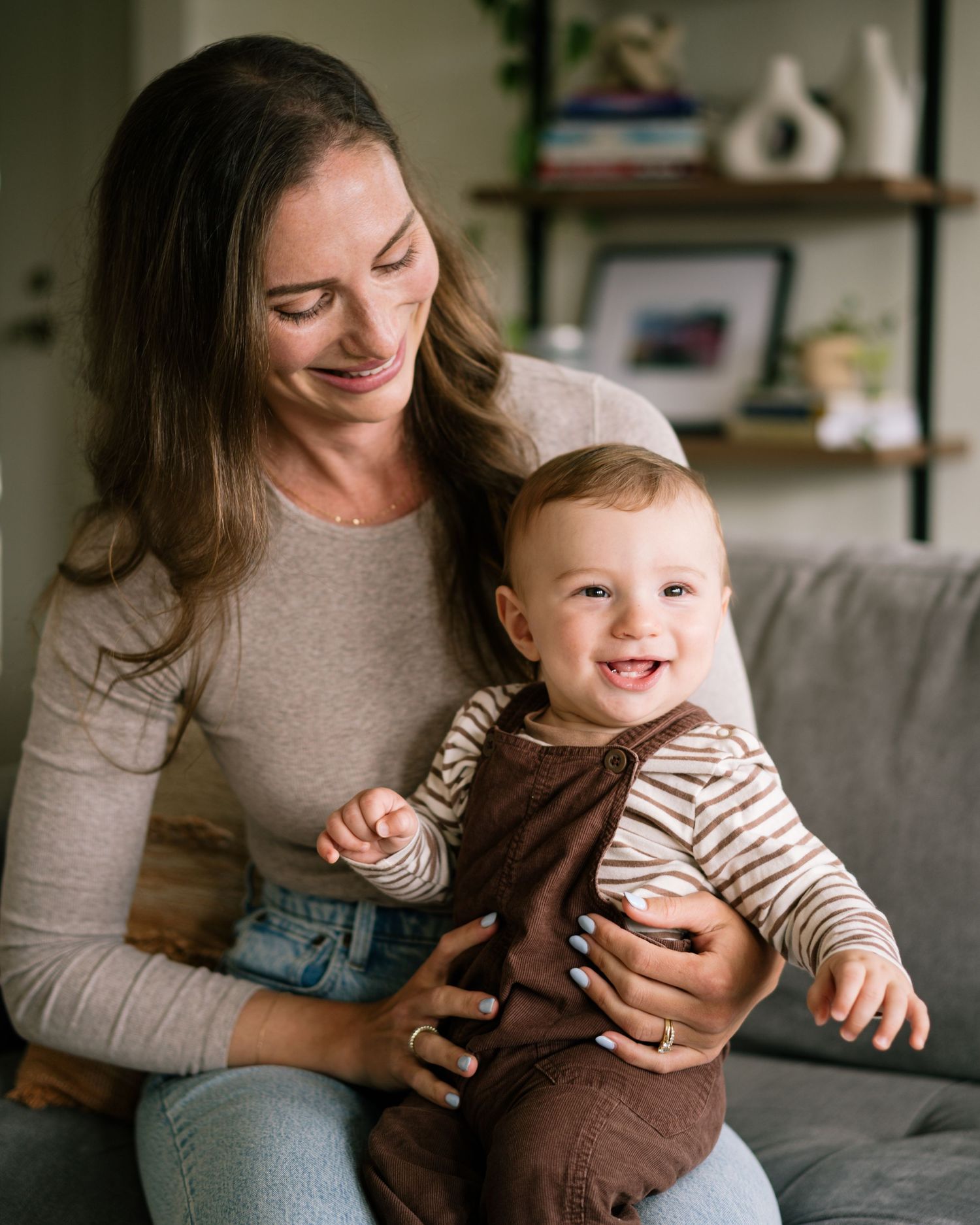 A smiling mother holds her happy baby in brown overalls on a cozy gray sofa indoors.